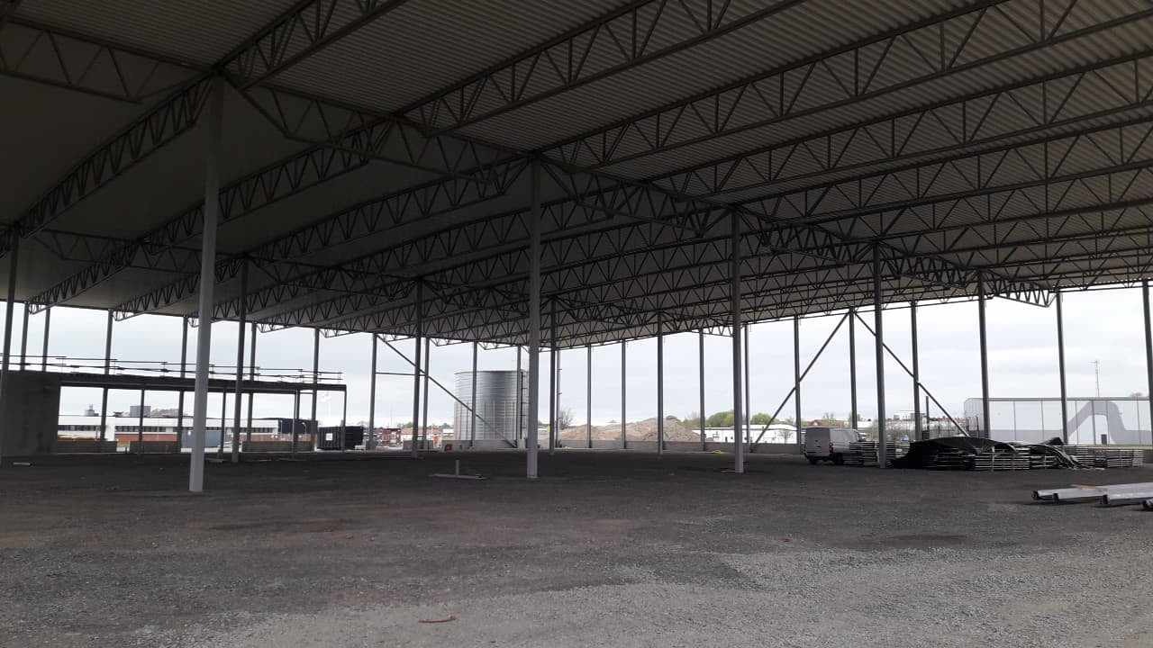 Wnętrze budowanej hali stalowej z widoczną konstrukcją dachu, słupami nośnymi i przygotowanym podłożem z kruszywa. Na zewnątrz widoczny silos i budynki.