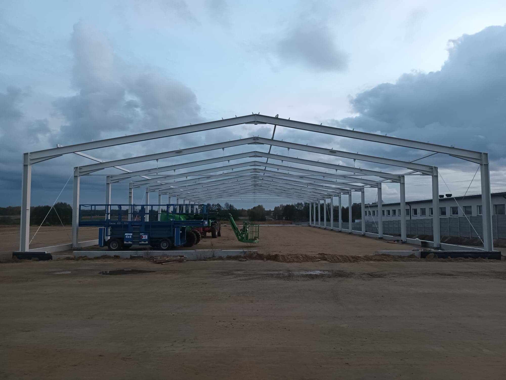 Stalowa konstrukcja hali w trakcie budowy, widoczne maszyny budowlane i pochmurne niebo.