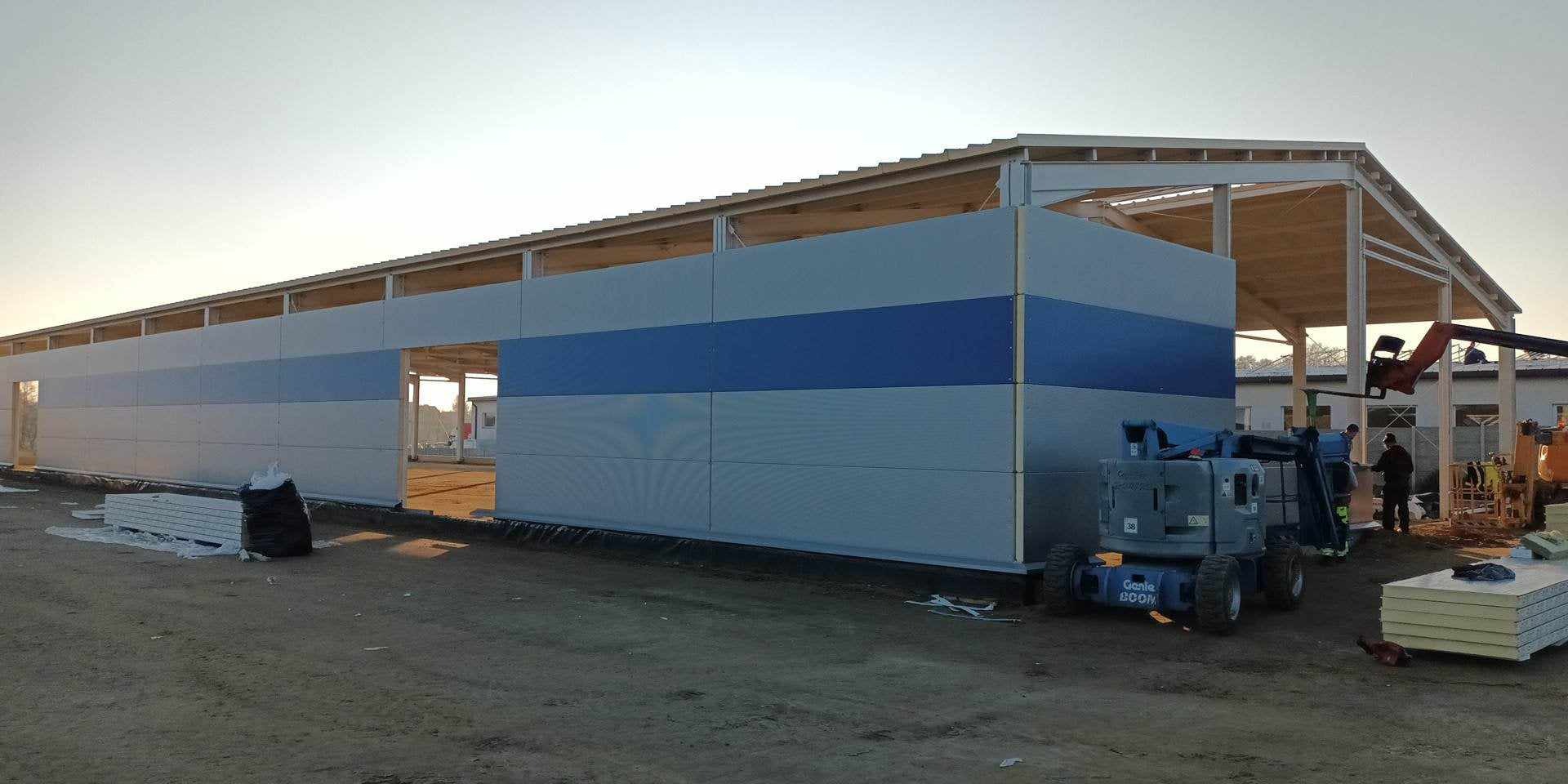 Budowa hali z płyt warstwowych w trakcie montażu, widoczny podnośnik Genie Boom i częściowo zamontowane ściany z niebieskim pasem, konstrukcja dachu w tle.