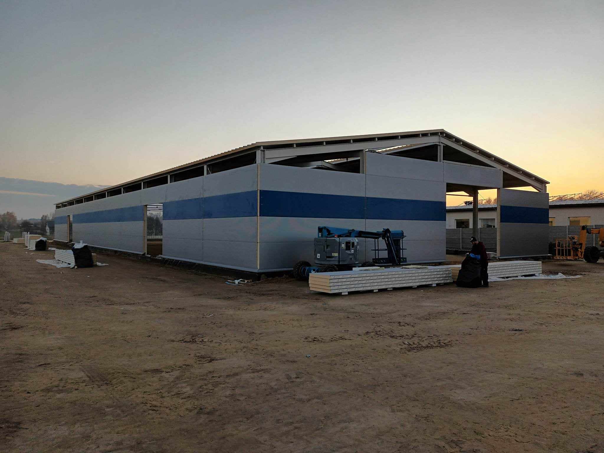 Wznoszona hala z szarych płyt z niebieskim pasem, widoczny podnośnik koszowy i ułożone płyty warstwowe, zachodzące słońce w tle.