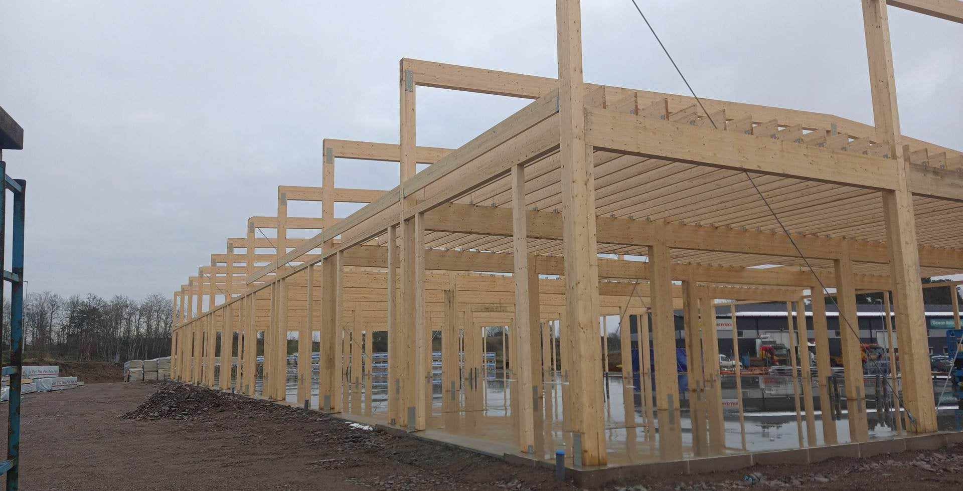 Wznoszenie drewnianej konstrukcji hali z widocznymi słupami, belkami i złączami, w tle stos płyt i teren budowy po deszczu.