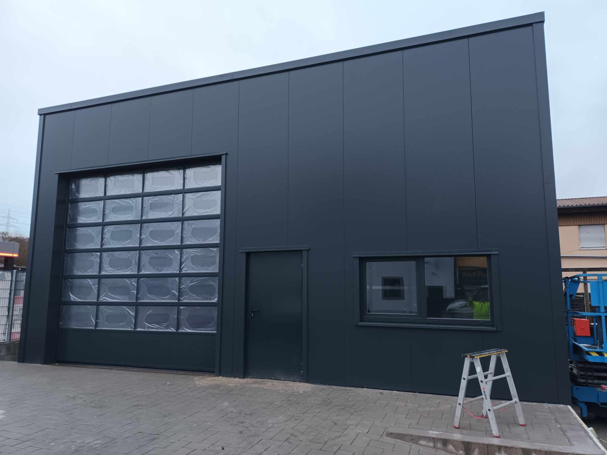 Nowo wybudowana hala z ciemnoszarej płyty warstwowej z bramą segmentową w folii ochronnej, widoczne okno i drzwi wejściowe, drabina aluminiowa oparta o ścianę, w tle podnośnik koszowy.