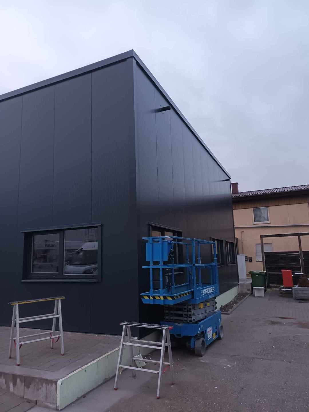 Nowoczesna hala przemysłowa z ciemnych płyt warstwowych, widoczny podnośnik koszowy i drabiny aluminiowe, w tle budynek mieszkalny.
