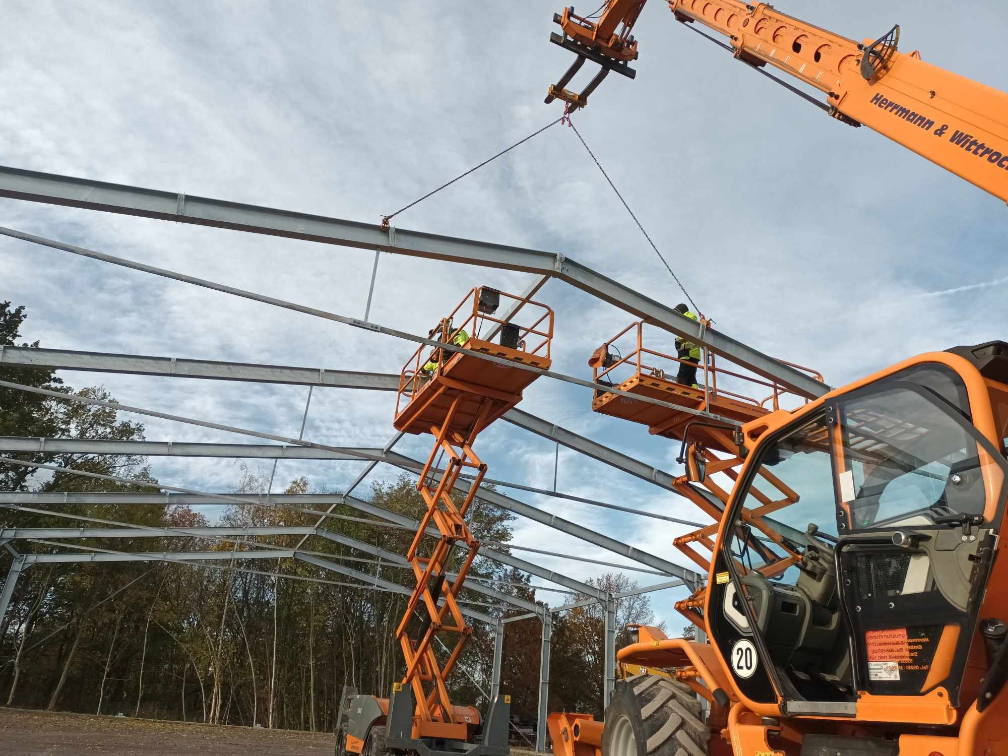 Montaż stalowej konstrukcji hali z wykorzystaniem dwóch podnośników nożycowych i dźwigu, widoczne elementy szkieletu hali na tle drzew i zachmurzonego nieba.