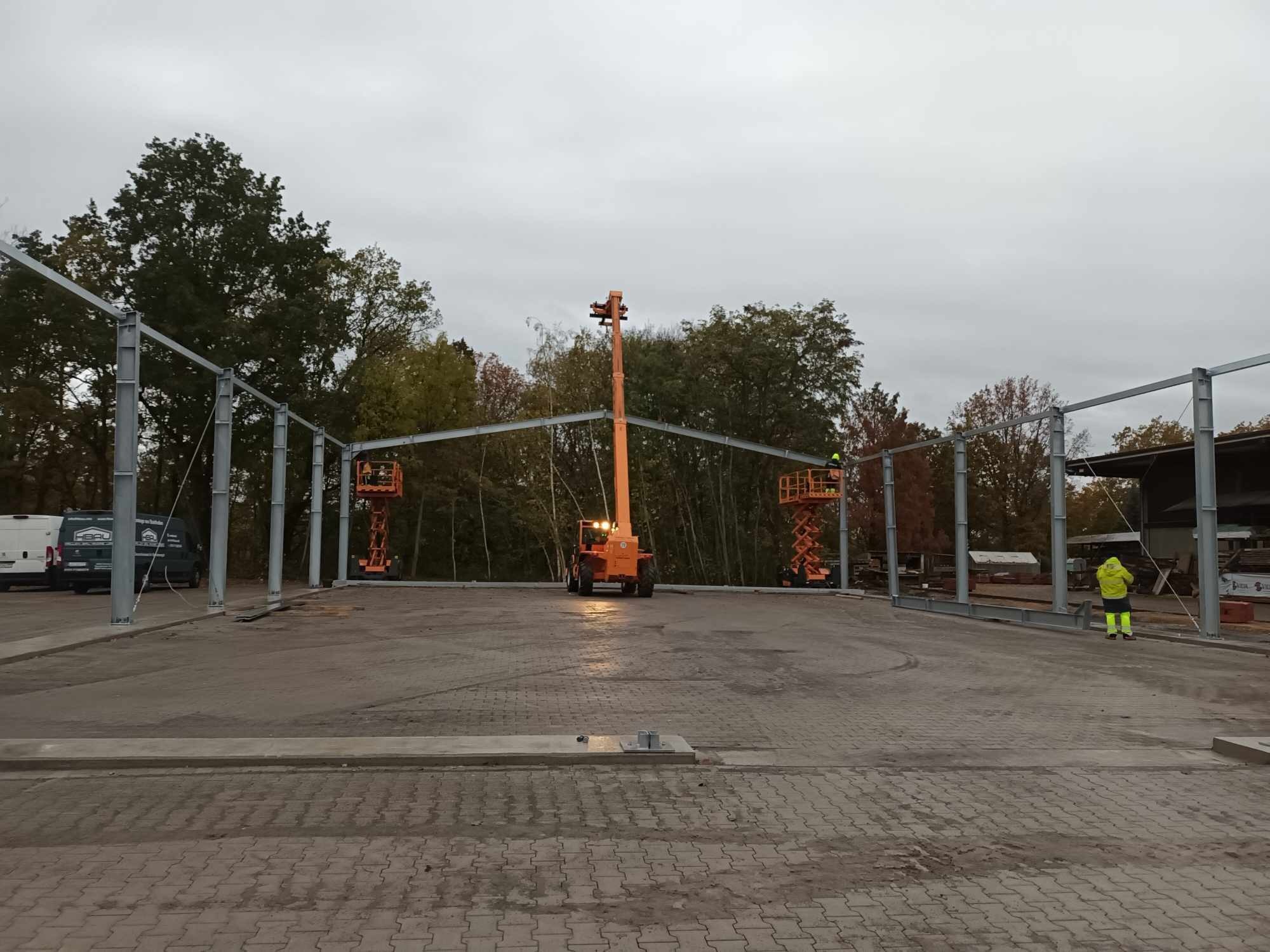 Stalowa konstrukcja hali w trakcie budowy, widoczne podnośniki koszowe i pracownik w żółtej kurtce, pochmurne niebo w tle.