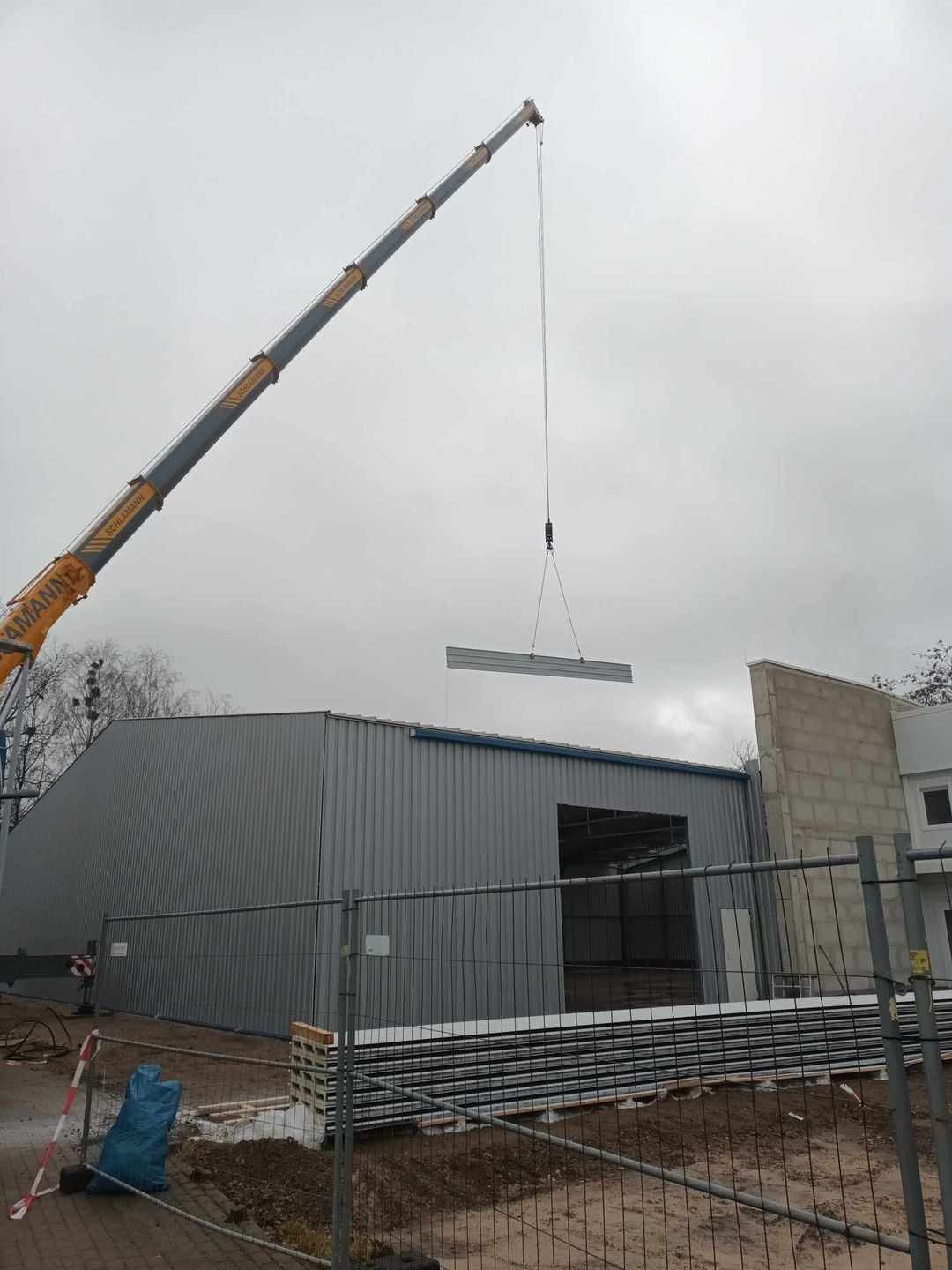 Montaż płyty warstwowej na hali stalowej za pomocą dźwigu Schlamann, widoczne zabezpieczenia budowy i stos płyt.