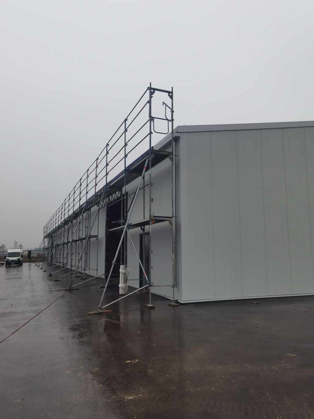 Długa hala w trakcie budowy, z widocznymi rusztowaniami wzdłuż ściany wykonanej z jasnych płyt. Na mokrym placu budowy stoi biały bus.