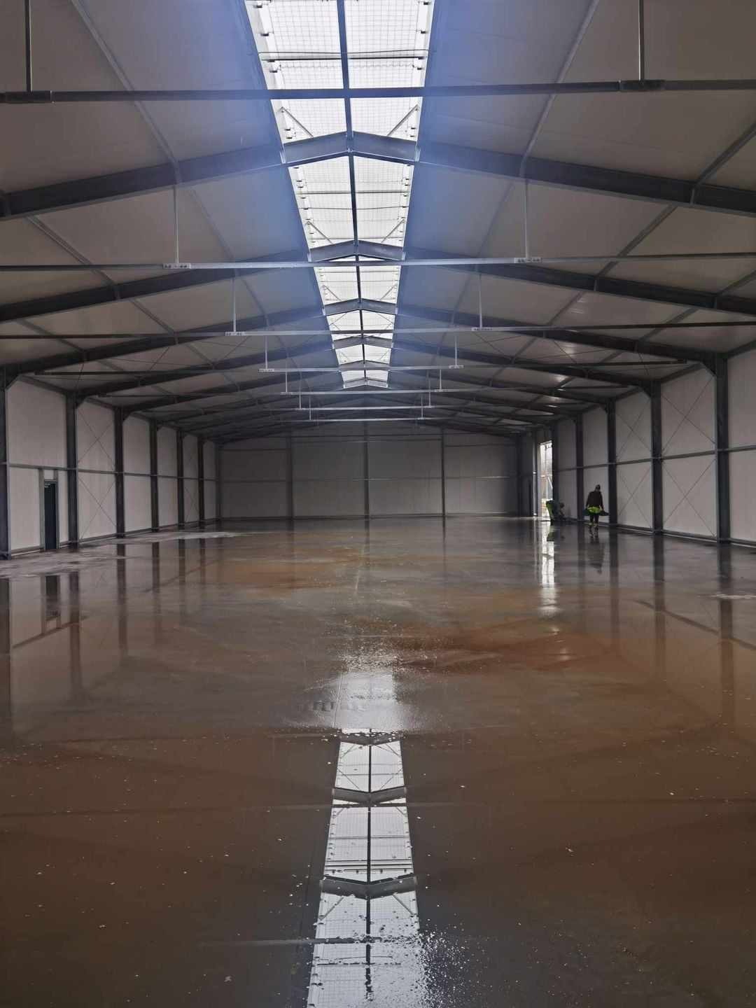 Wnętrze nowej hali przemysłowej z widoczną konstrukcją stalową, panelami ściennymi i mokrą posadzką odbijającą świetliki dachowe; dwóch pracowników w oddali.