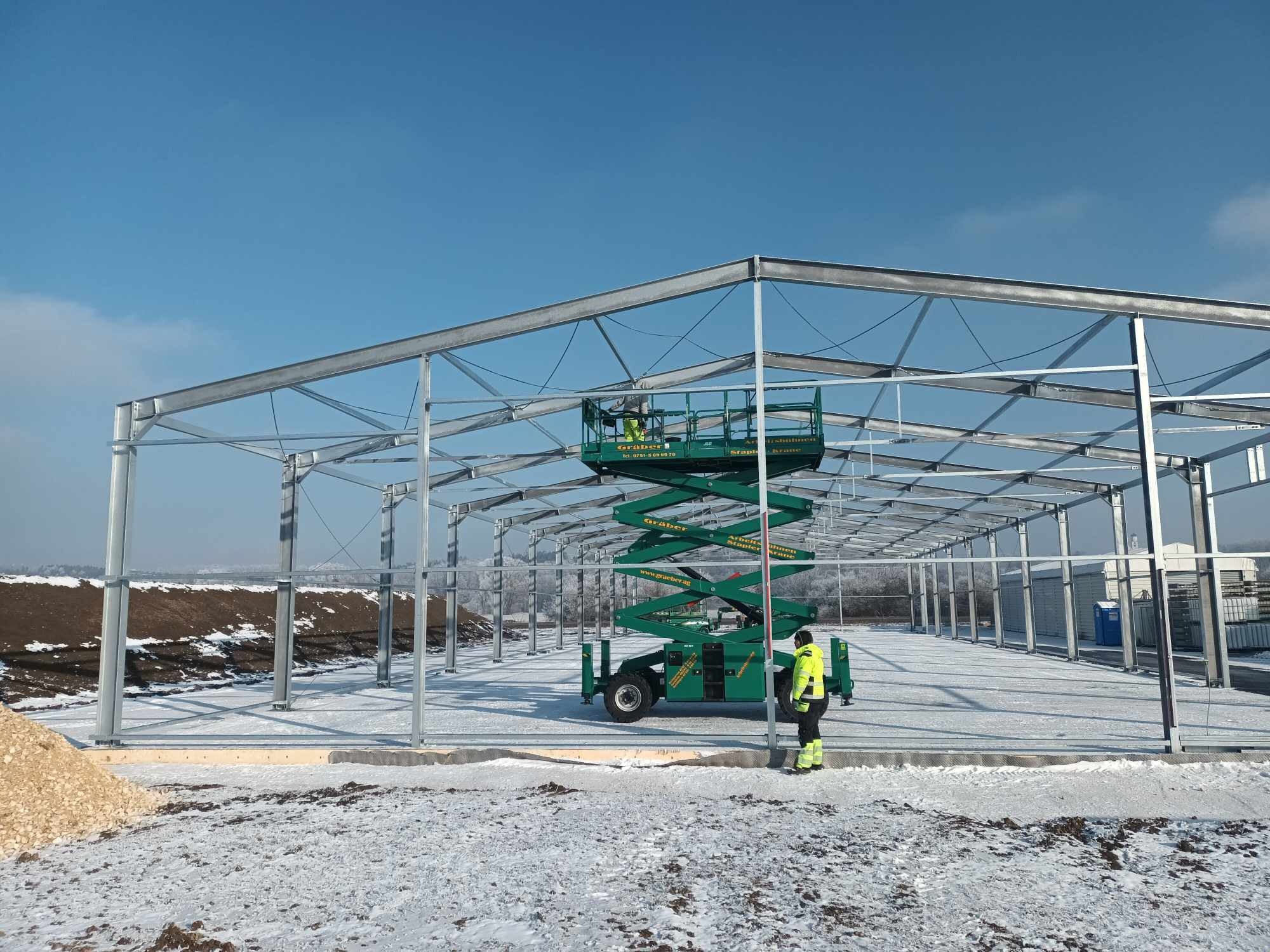 Stalowa konstrukcja hali w trakcie budowy, widoczny podnośnik nożycowy z pracownikami na platformie, śnieg na ziemi, błękitne niebo.