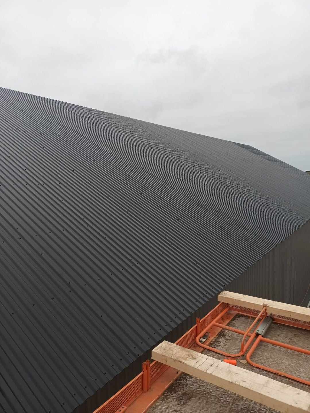 Widok z góry na fragment dachu hali przemysłowej pokrytego ciemnoszarą blachą trapezową, z pomarańczowym rusztowaniem na pierwszym planie, pochmurne niebo w tle.
