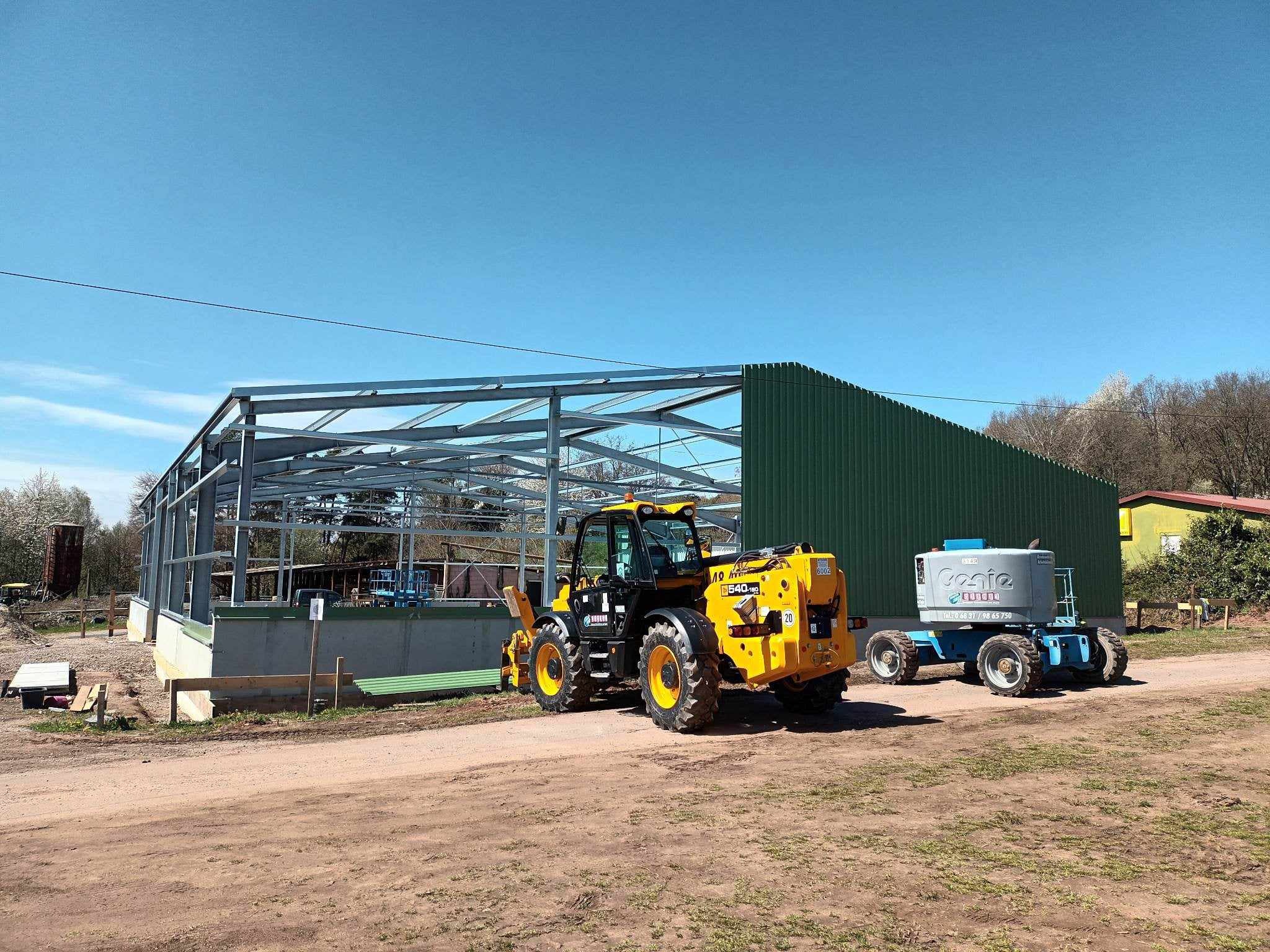 Wznoszona hala z częściowo zamontowaną zieloną ścianą z płyt warstwowych, na pierwszym planie żółta ładowarka teleskopowa i podnośnik koszowy na ziemnym placu budowy w słoneczny dzień.