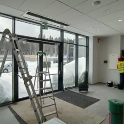 Pracownik w odblaskowej kamizelce czyści framugę drzwi w nowoczesnym budynku, w tle widoczne drabiny opierające się o przeszkloną ścianę z widokiem na zasypany śniegiem teren.