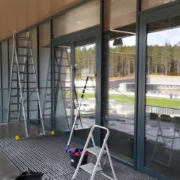 Dwie drabiny i sprzęt do mycia okien ustawione przed dużymi oknami z widokiem na las w trakcie prac porządkowych.