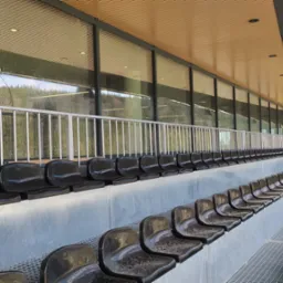 Rzędy czarnych, mokrych siedzeń na trybunach stadionu, widoczne krople wody po deszczu, w tle szklana ściana z odbiciem drzew i nieba.