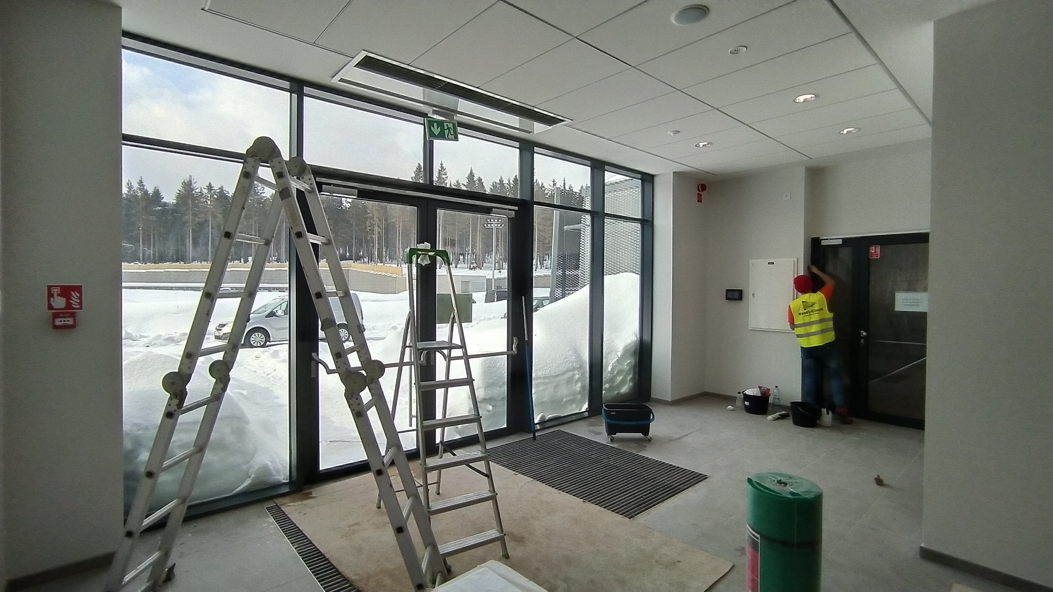 Pracownik w odblaskowej kamizelce czyści framugę drzwi w nowoczesnym budynku, w tle widoczne drabiny opierające się o przeszkloną ścianę z widokiem na zasypany śniegiem teren.