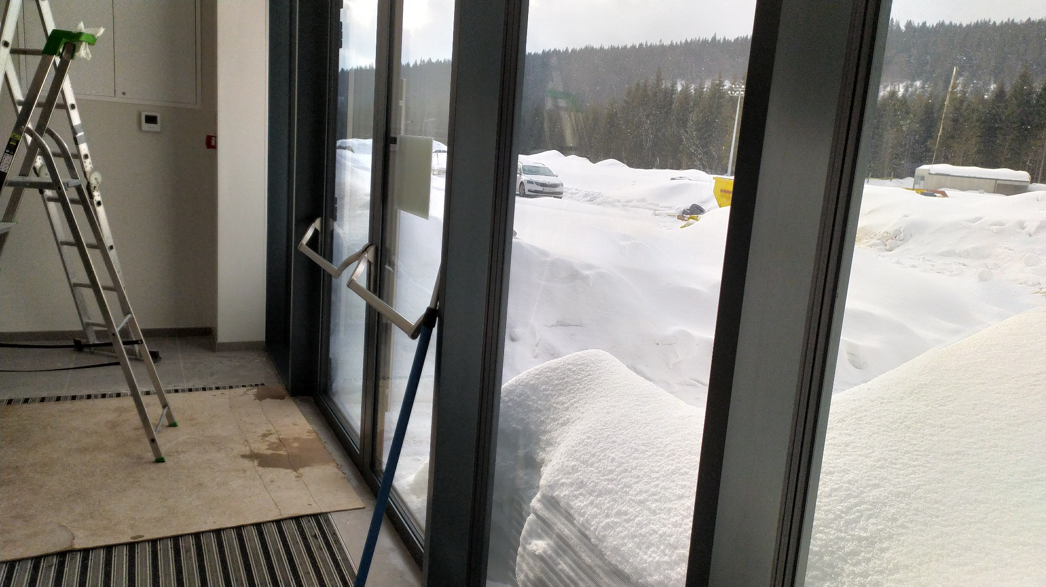 Wnętrze budynku z widokiem na zasypany śniegiem teren przez duże okna; drabina oparta o ścianę, mokra plama na podłodze przy wejściu.