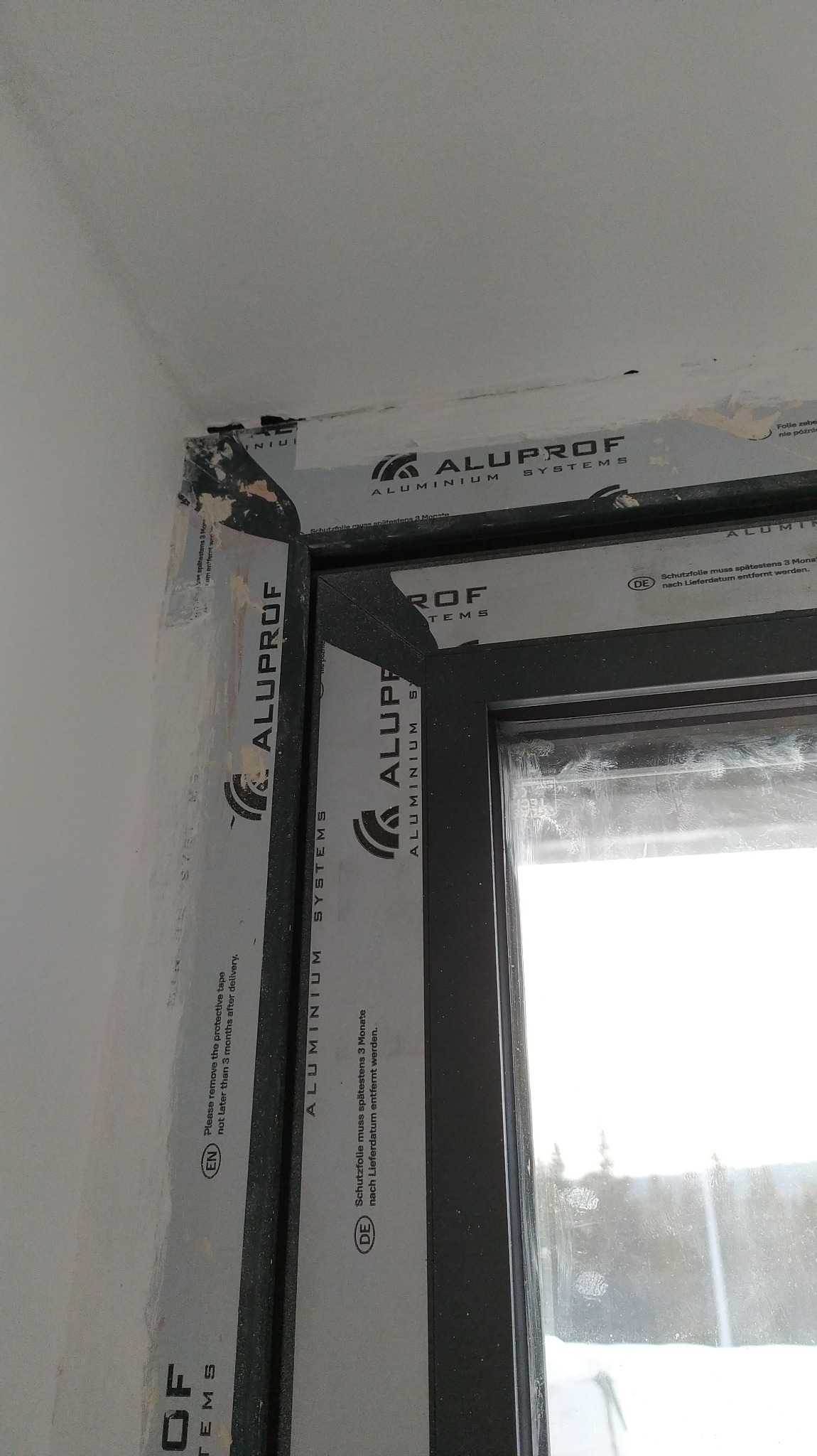 Fragment okna z czarną ramą Aluplast, częściowo usunięta folia ochronna z profili, widoczne ślady po remoncie na ścianie i suficie.