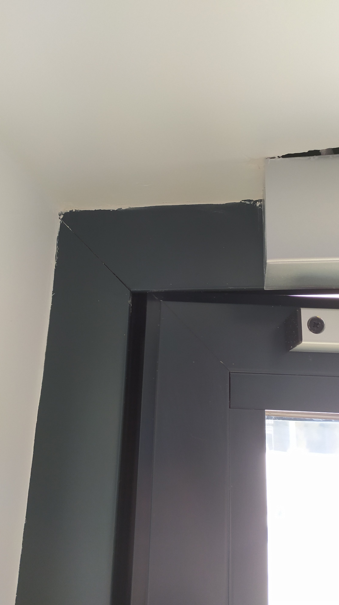 Niedokładnie pomalowana, ciemnoszara rama okienna z widocznymi śladami farby na ścianie i suficie.