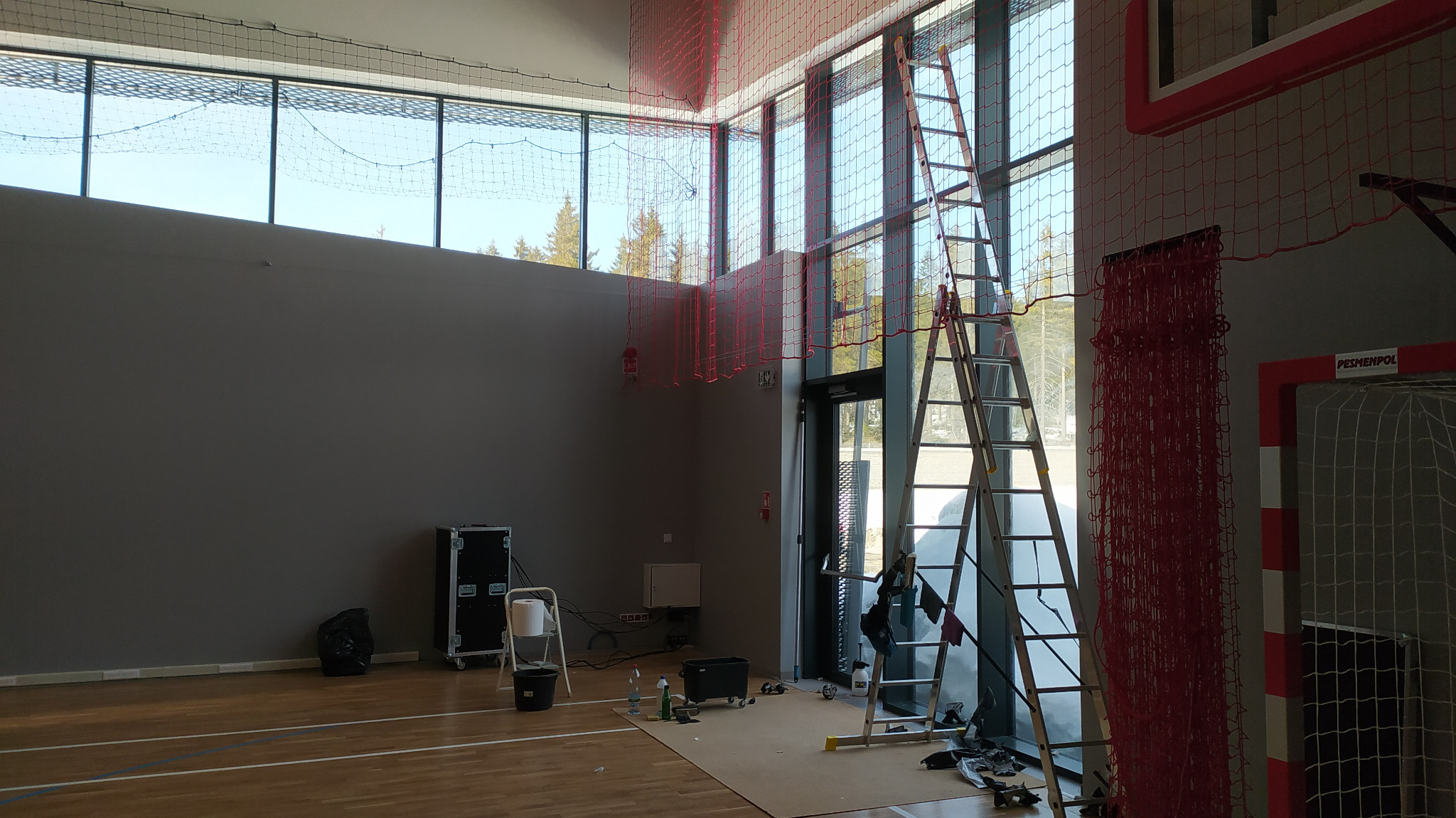 Wysoka drabina oparta o okna w hali sportowej, widoczne akcesoria do mycia i sprzątania, siatka zabezpieczająca pod sufitem.