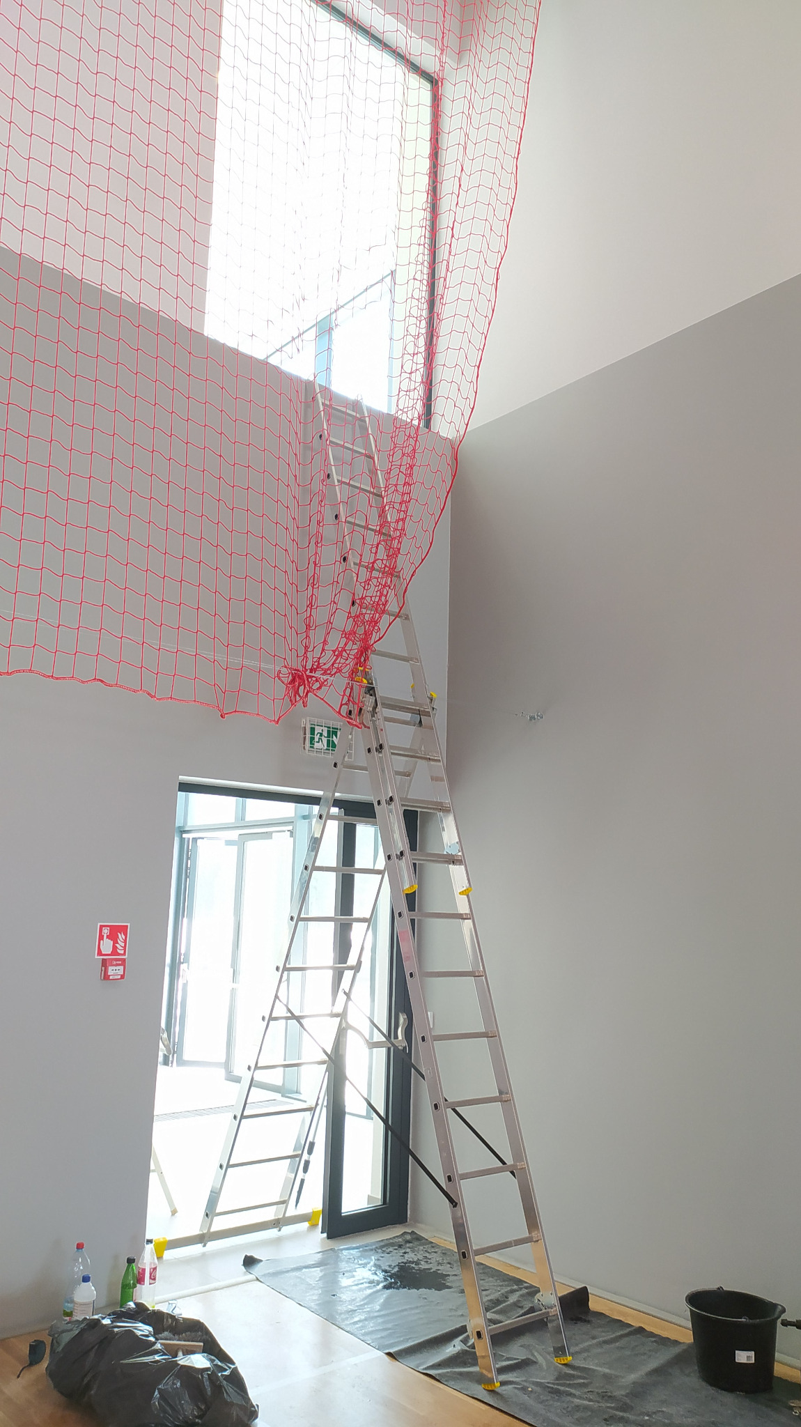 Wysoka drabina aluminiowa oparta o okno zabezpieczone czerwoną siatką ochronną w surowym, jasnym pomieszczeniu, podłoga zabezpieczona folią malarską, widoczne butelki z płynami i czarne wiadro.