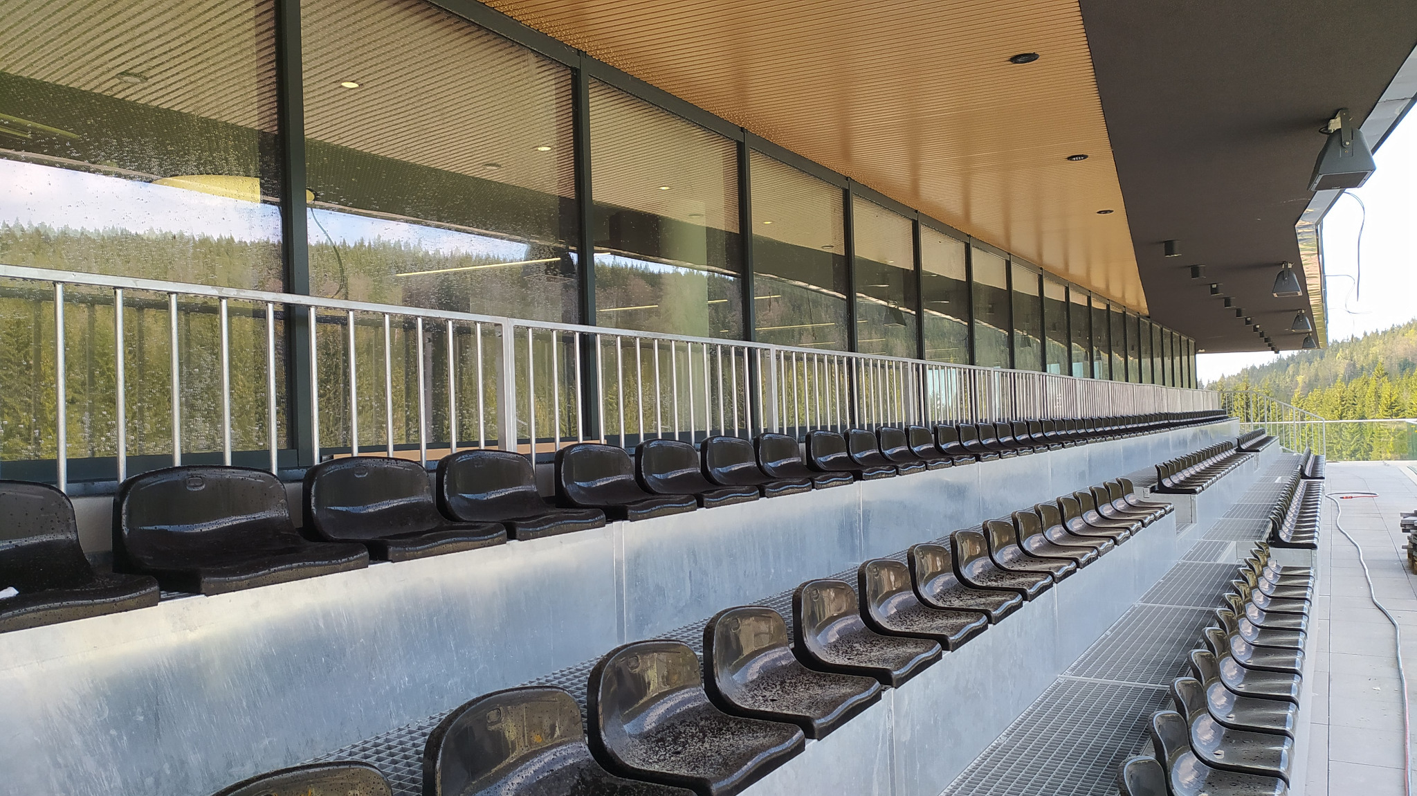 Rzędy czarnych, mokrych siedzeń na trybunach stadionu, widoczne krople wody po deszczu, w tle szklana ściana z odbiciem drzew i nieba.