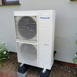 Zewnętrzna jednostka klimatyzacyjna Panasonic, umieszczona na betonowych podstawach, przy szarej ścianie budynku z widocznym oknem i fragmentem ceglanego chodnika.