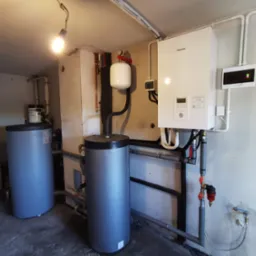 Instalacja grzewcza w Trzebnicy: biały piec gazowy Panasonic, dwa szare zbiorniki akumulacyjne, rury i armatura w piwnicy z betonową podłogą.