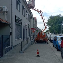 Piotr Obst - Pomarańczowy podnośnik koszowy na chodniku przed budynkiem z lokalami usługowymi. Widoczny stożek ostrzegawczy i zaparkowane samochody. Dzień, pochmurne niebo.