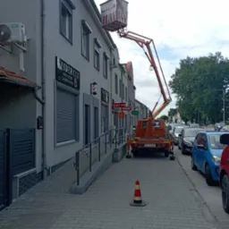 Pomarańczowy podnośnik koszowy na chodniku przed budynkiem z lokalami usługowymi. Widoczny stożek ostrzegawczy i zaparkowane samochody. Dzień, pochmurne niebo.