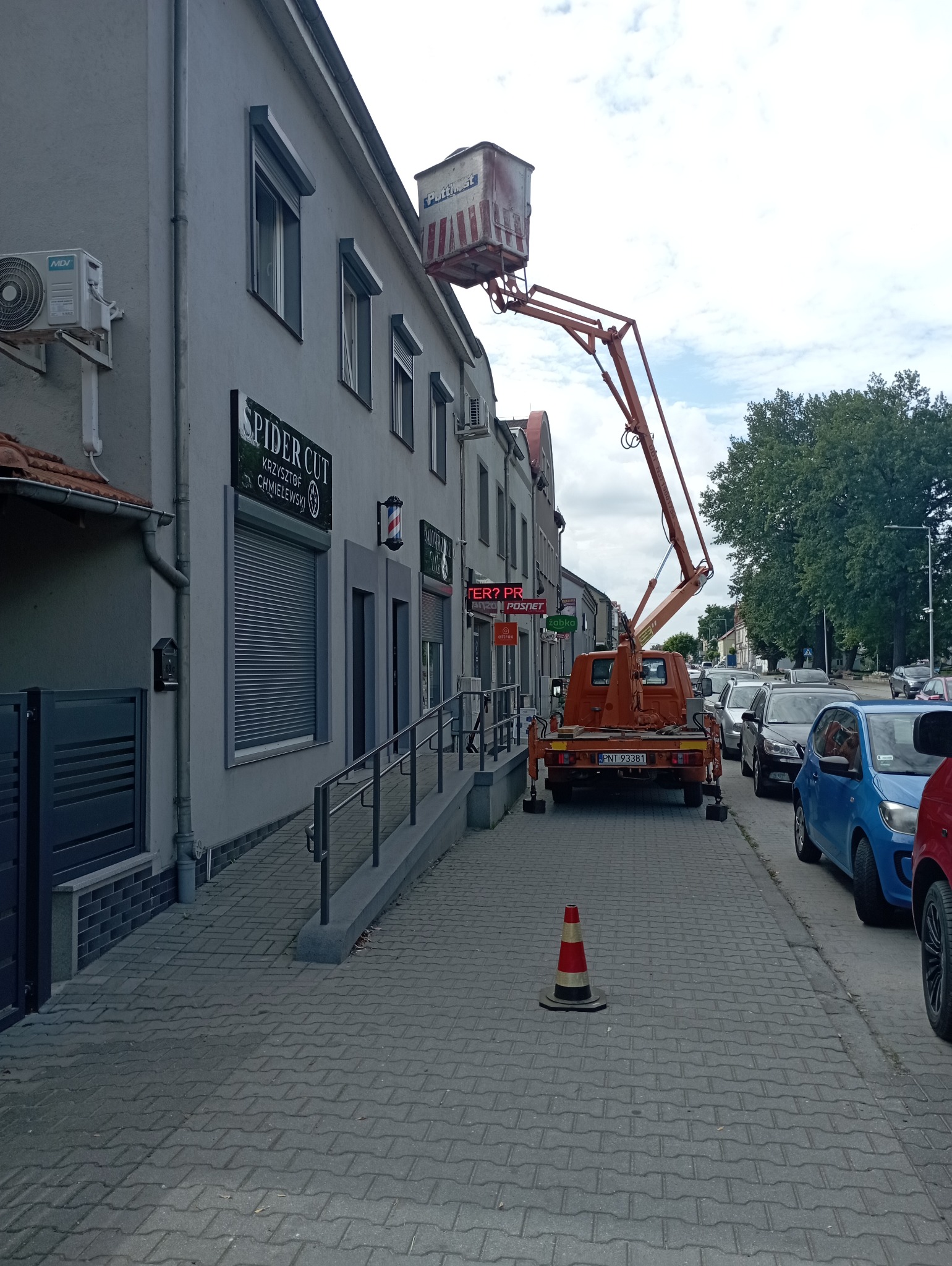 Pomarańczowy podnośnik koszowy na chodniku przed budynkiem z lokalami usługowymi. Widoczny stożek ostrzegawczy i zaparkowane samochody. Dzień, pochmurne niebo.