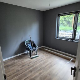 Remonty Adrian Stypuła - Pokój po remoncie: brązowe panele podłogowe, szare ściany, plastikowe krzesło z miotłą, okno z widokiem na zieleń. Wykończenie wnętrza, widok zza otwartych drzwi.