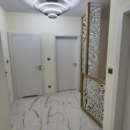 Remonty Adrian Stypuła - Jasne wnętrze przedpokoju z marmurową podłogą, białymi drzwiami i dekoracyjną, ażurową ścianką działową z drewnianą ramą, oświetlone nowoczesną lampą sufitową.