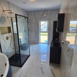 Remonty Adrian Stypuła - Elegancka łazienka z kabiną prysznicową typu walk-in, marmurowymi płytkami, czarną armaturą i szafką z umywalką. Widok na okno i dom w tle.