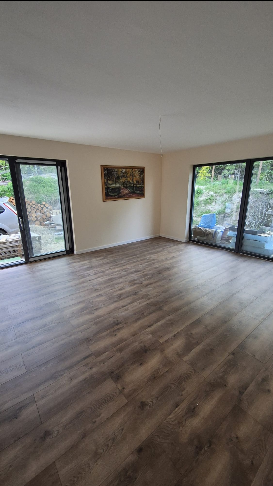 Jasne wnętrze z panelami podłogowymi w odcieniach brązu, duże okna z widokiem na zieleń, obraz na ścianie. Minimalistyczny wystrój.