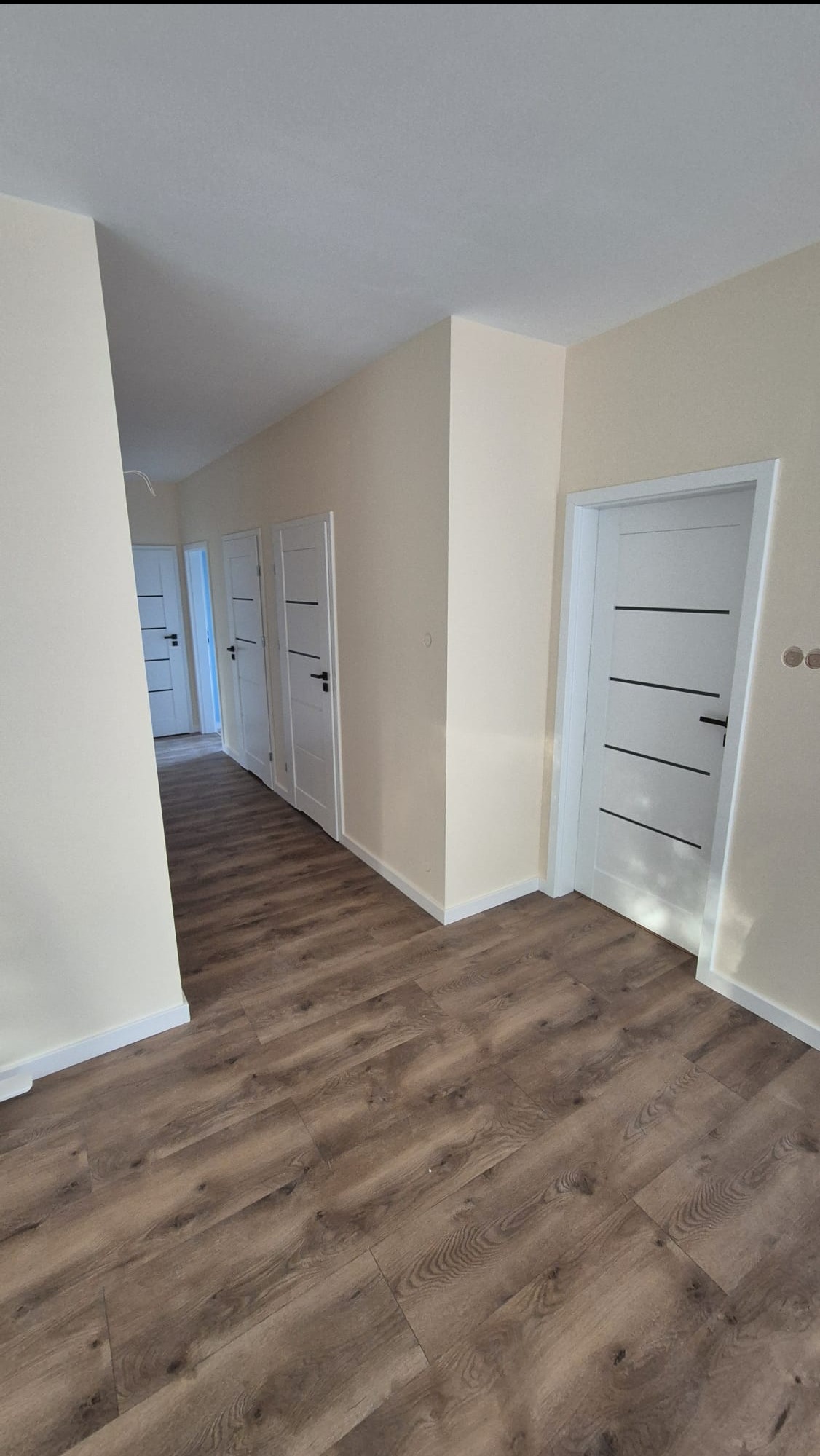 Jasne wnętrze po remoncie: brązowe panele podłogowe, kremowe ściany i białe, nowoczesne drzwi z czarnymi akcentami w perspektywie korytarza.