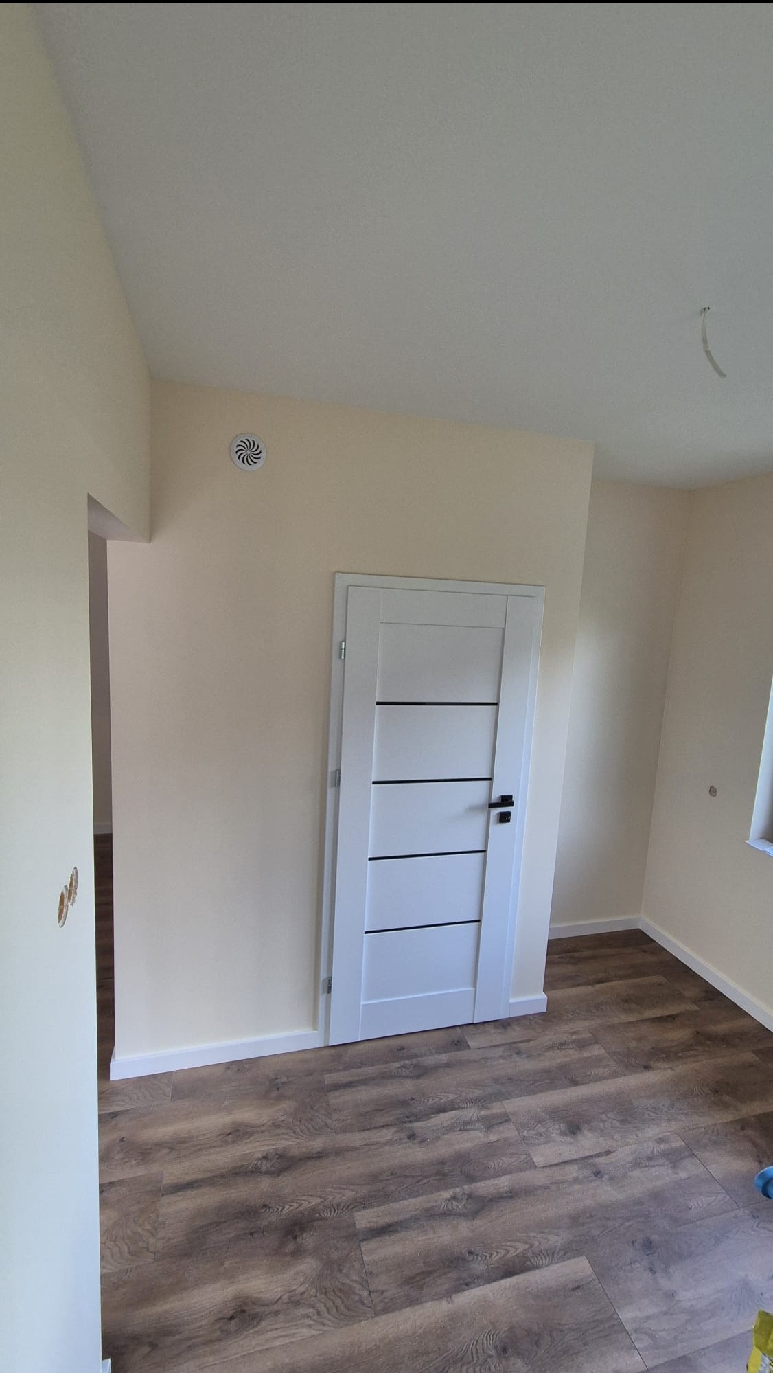 Wykończone wnętrze pokoju z panelami podłogowymi i białą, nowoczesną stolarką drzwiową. Ściany w odcieniach beżu, widoczny nawiewnik wentylacyjny.