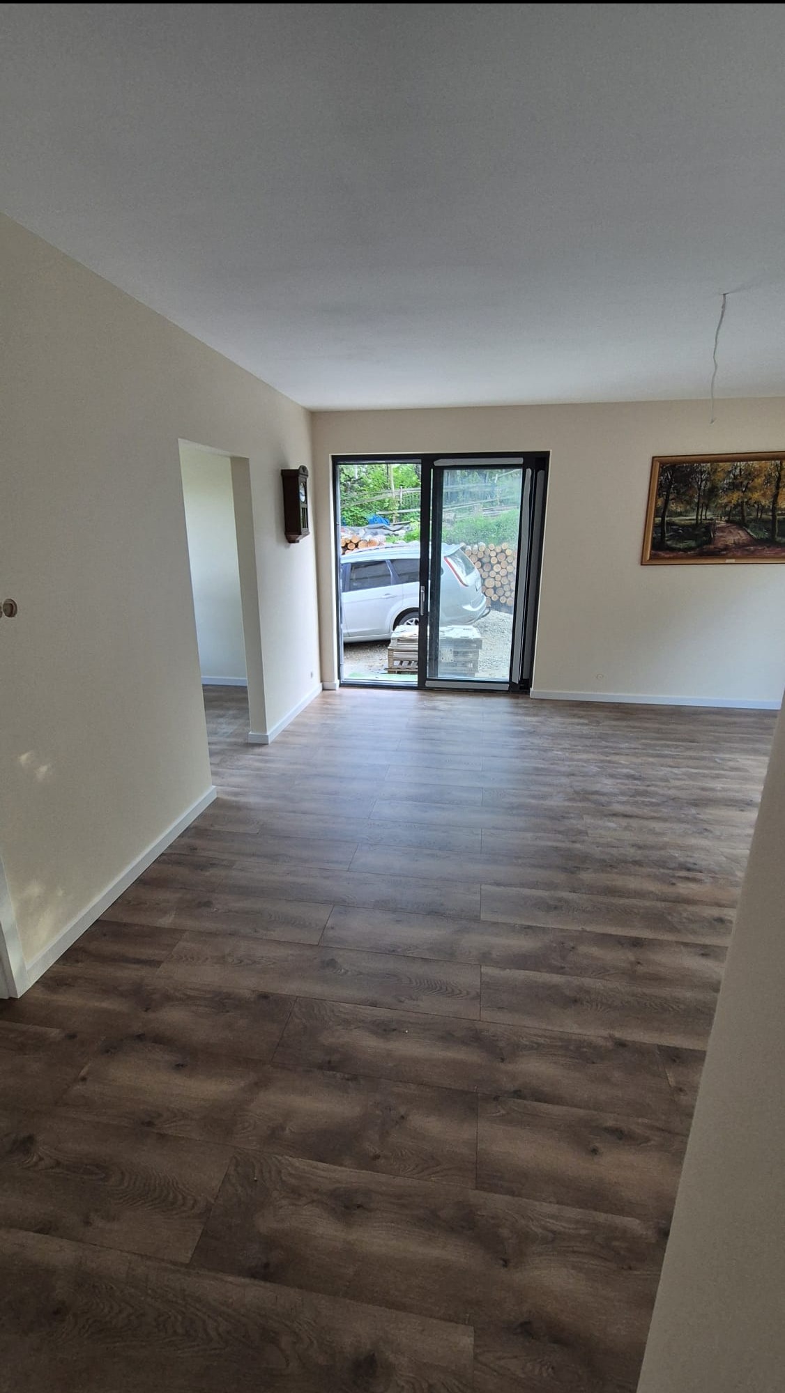 Wnętrze pokoju z panelami podłogowymi w odcieniach brązu. Widoczne ściany w jasnym kolorze, okno przesuwne z widokiem na zewnątrz i dekoracje ścienne.