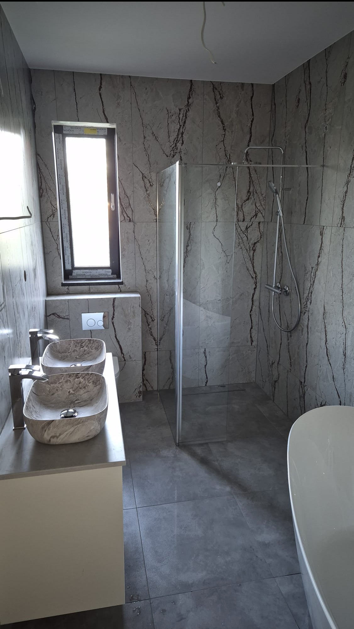 Nowoczesna łazienka z marmurowymi płytkami, kabiną prysznicową typu walk-in i podwójną umywalką na blacie. Minimalistyczne wyposażenie, szara kolorystyka, duże okno.