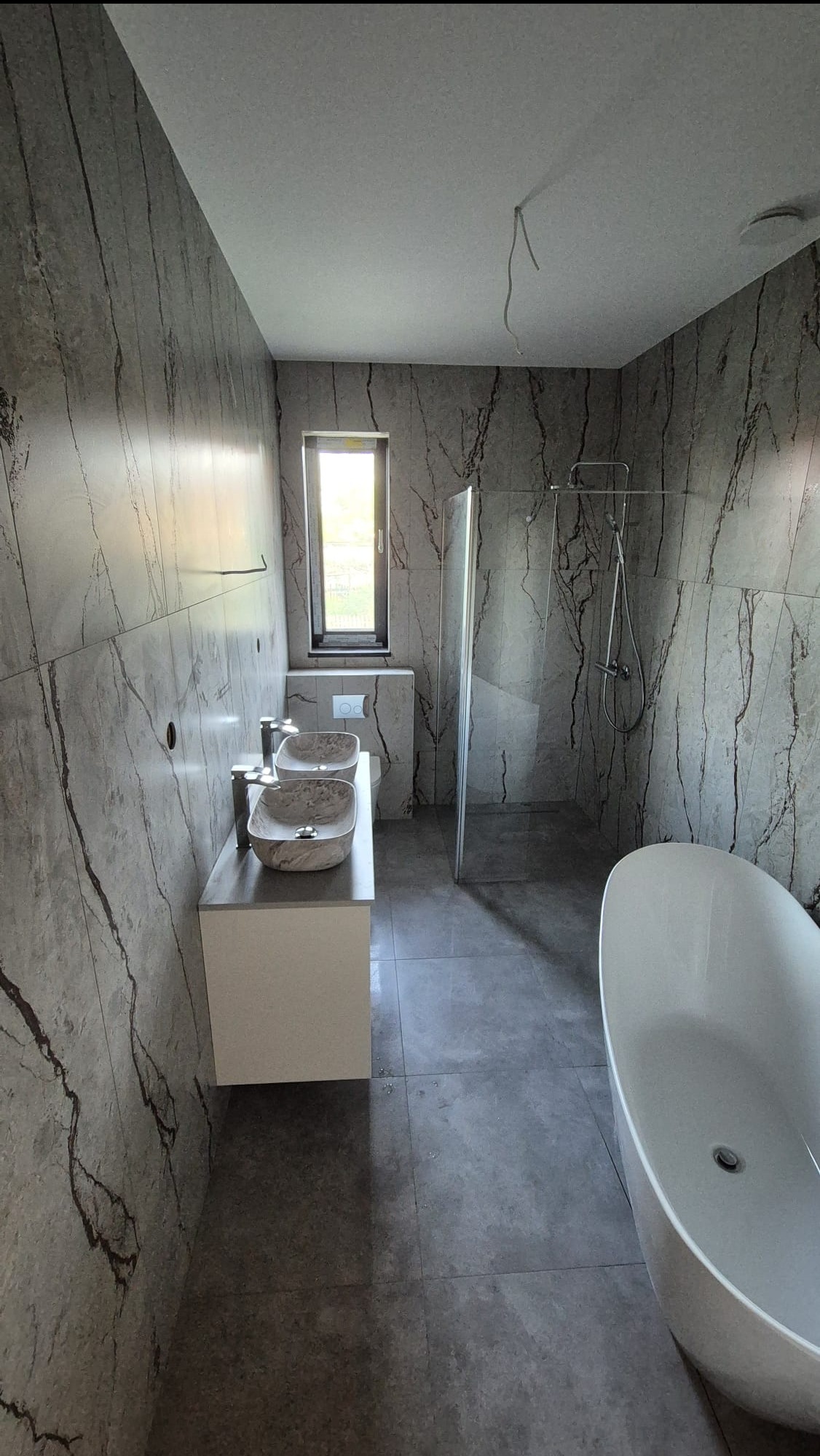 Nowoczesna łazienka z płytkami imitującymi marmur, owalną wanną i dwiema umywalkami w kształcie mis na blacie. Minimalistyczny design, szklana kabina prysznicowa.