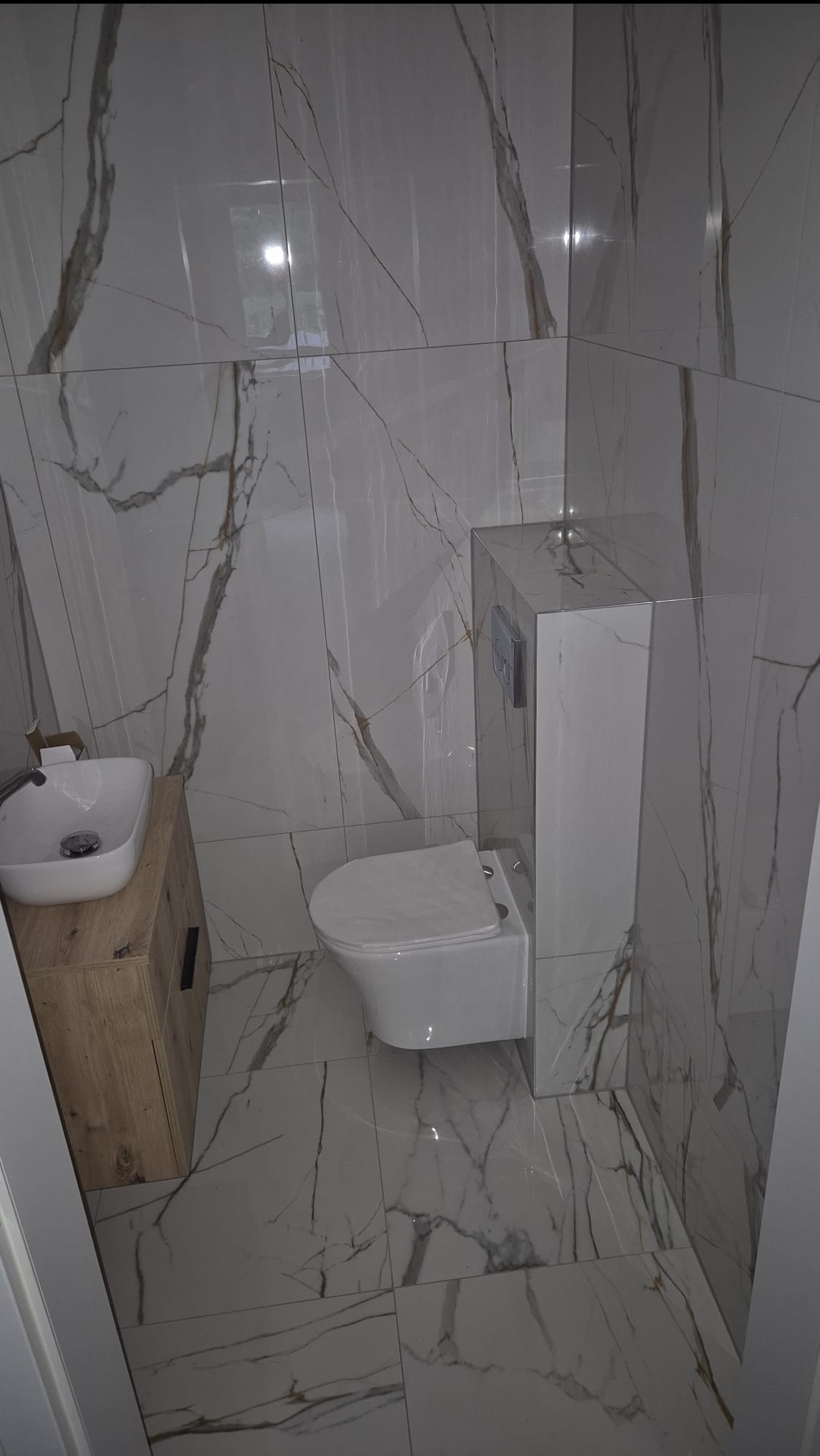 Nowoczesna łazienka z białymi płytkami imitującymi marmur, drewnianą szafką pod umywalką i podwieszaną toaletą. Minimalistyczny design, błyszczące wykończenie.