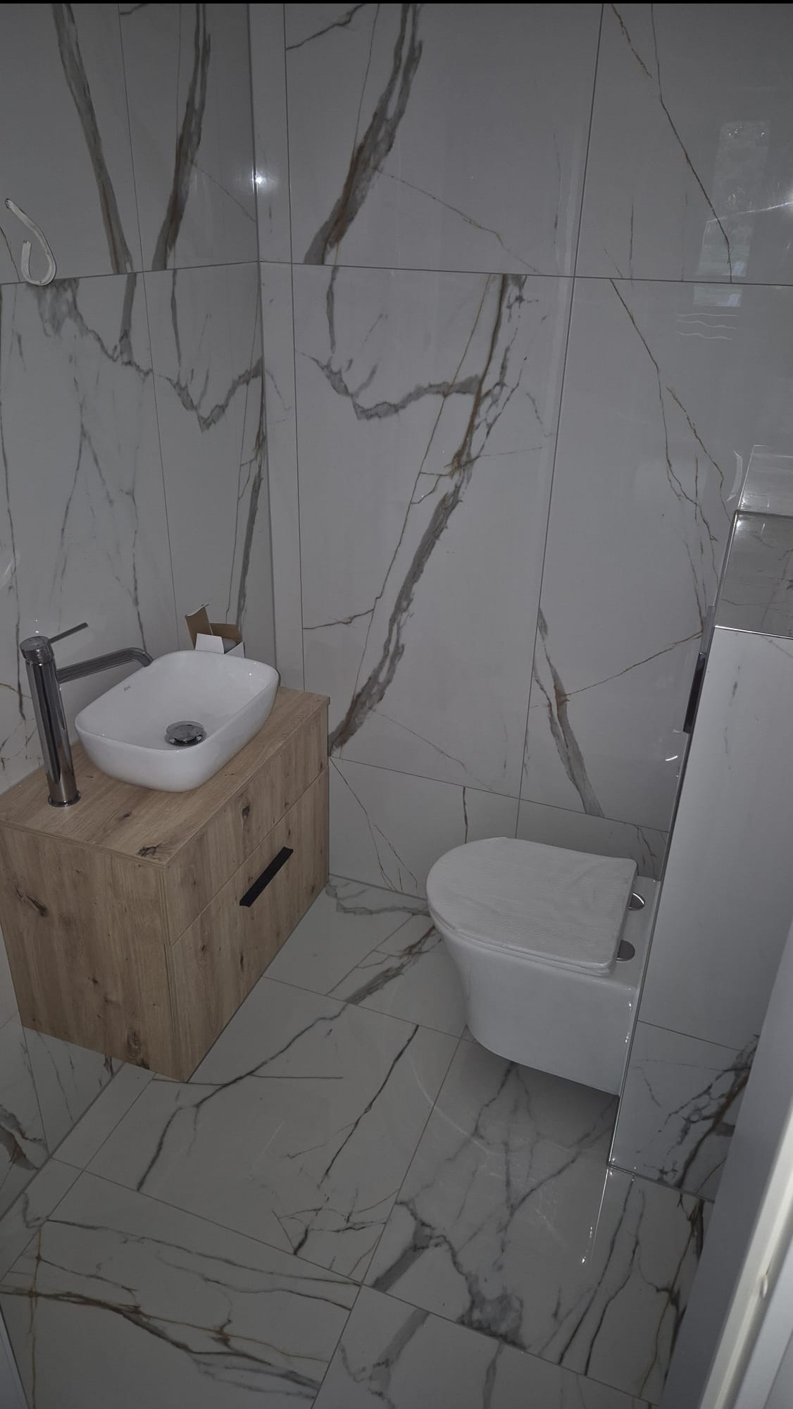 Nowoczesna łazienka po remoncie: białe płytki z marmurowym wzorem, drewniana szafka pod umywalką z chromowaną baterią i podwieszana toaleta.