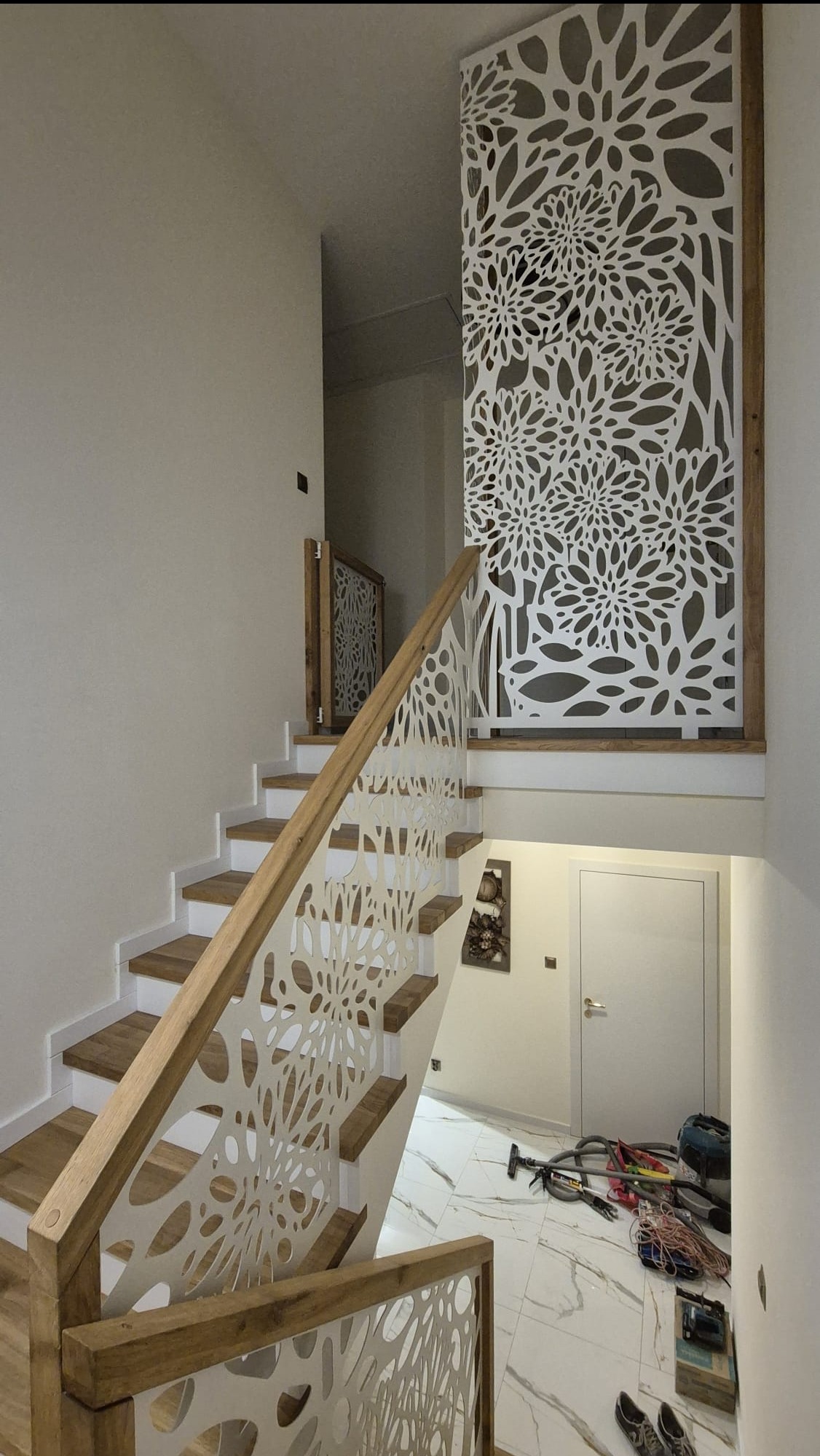 Schody z drewnianymi stopniami i ażurową, białą balustradą z motywem kwiatowym, prowadzące na górę. Na podłodze widoczne narzędzia i akcesoria.