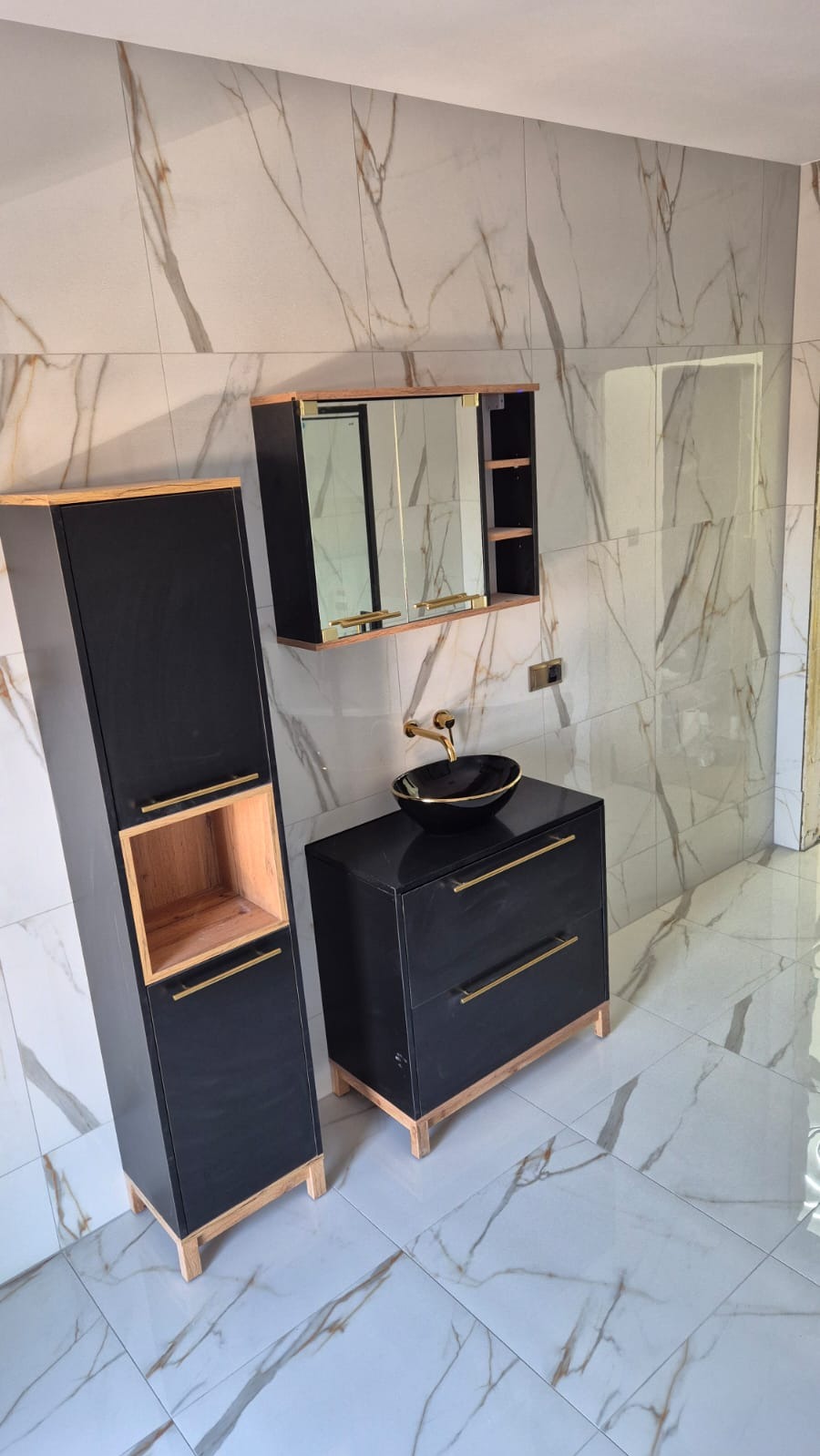 Nowoczesna łazienka z czarnymi szafkami i umywalką na tle marmurowych płytek ściennych i podłogowych. Złote akcenty podkreślają luksusowy charakter.