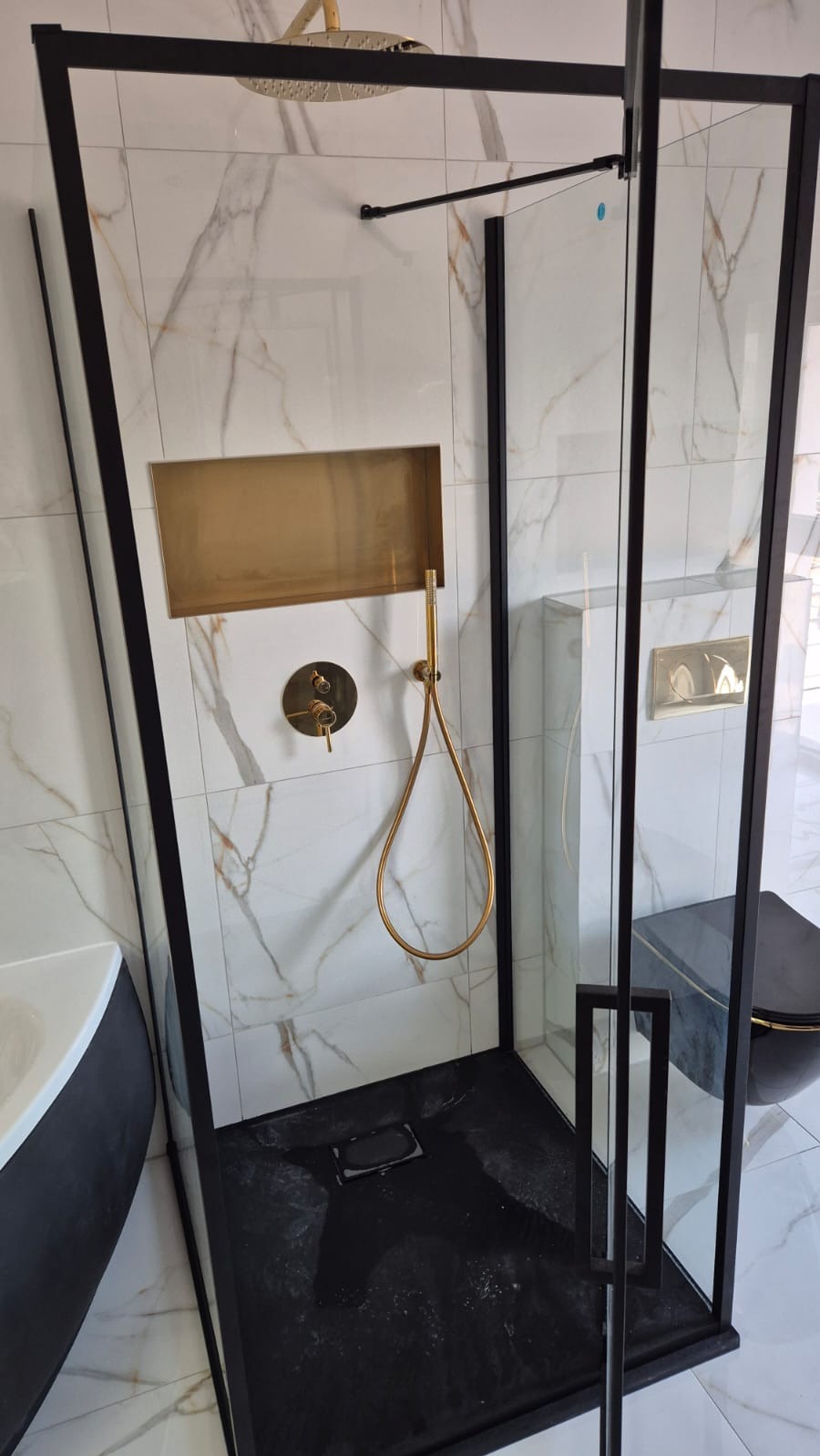 Nowoczesna kabina prysznicowa z czarną ramą i złotymi akcentami, marmurowe płytki na ścianach, czarny brodzik. Widoczny fragment wanny i toalety w tym samym stylu.