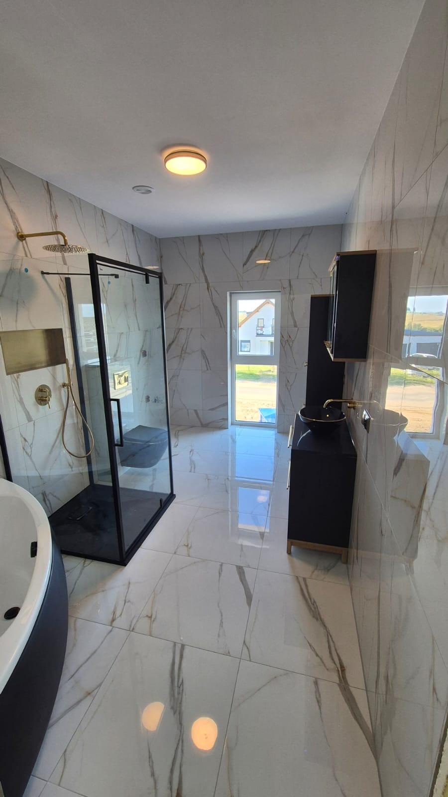Elegancka łazienka z kabiną prysznicową typu walk-in, marmurowymi płytkami, czarną armaturą i szafką z umywalką. Widok na okno i dom w tle.