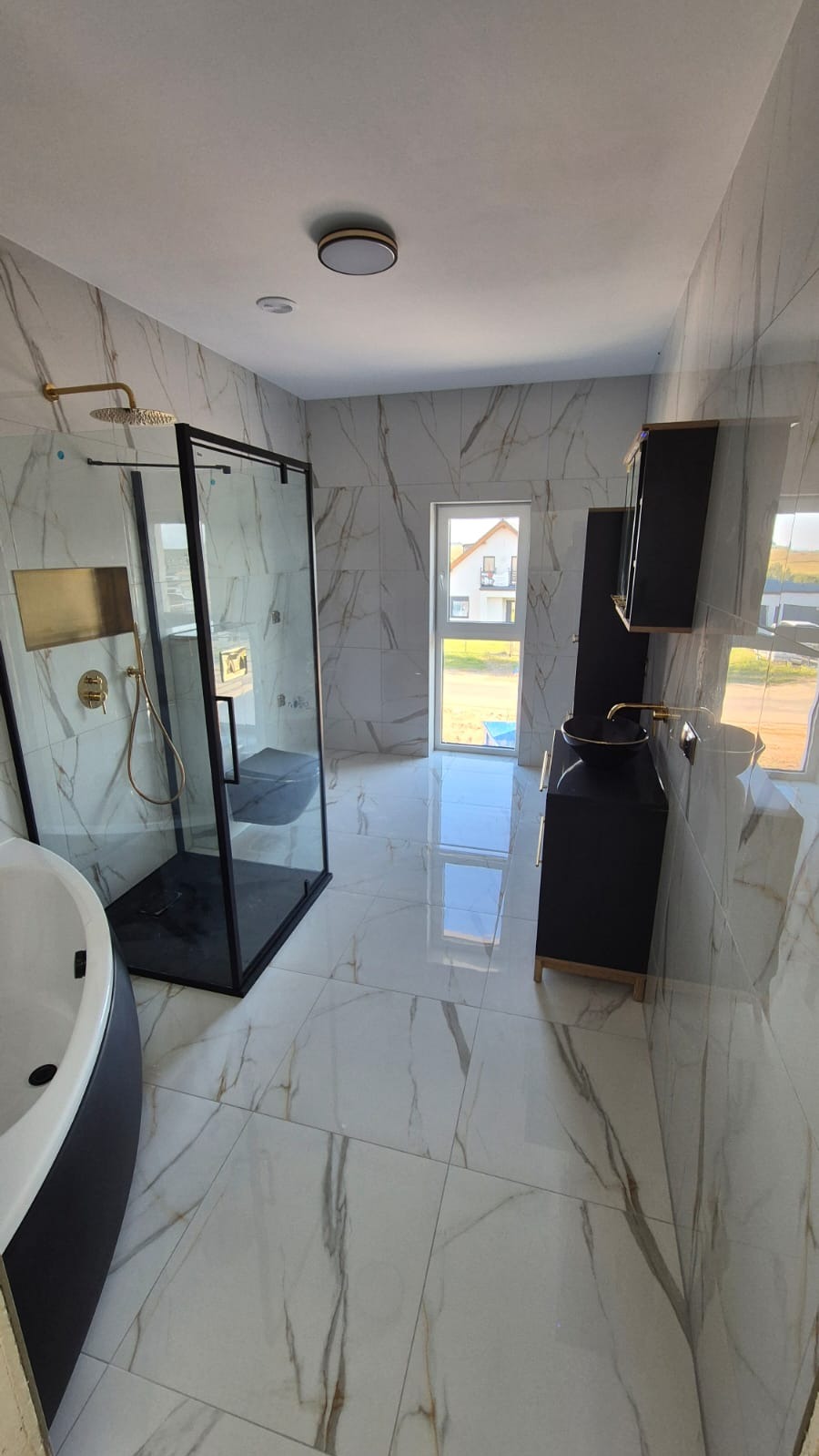 Nowoczesna łazienka z czarną kabiną prysznicową, wanną i szafkami na tle białych płytek imitujących marmur. Złote detale armatury.