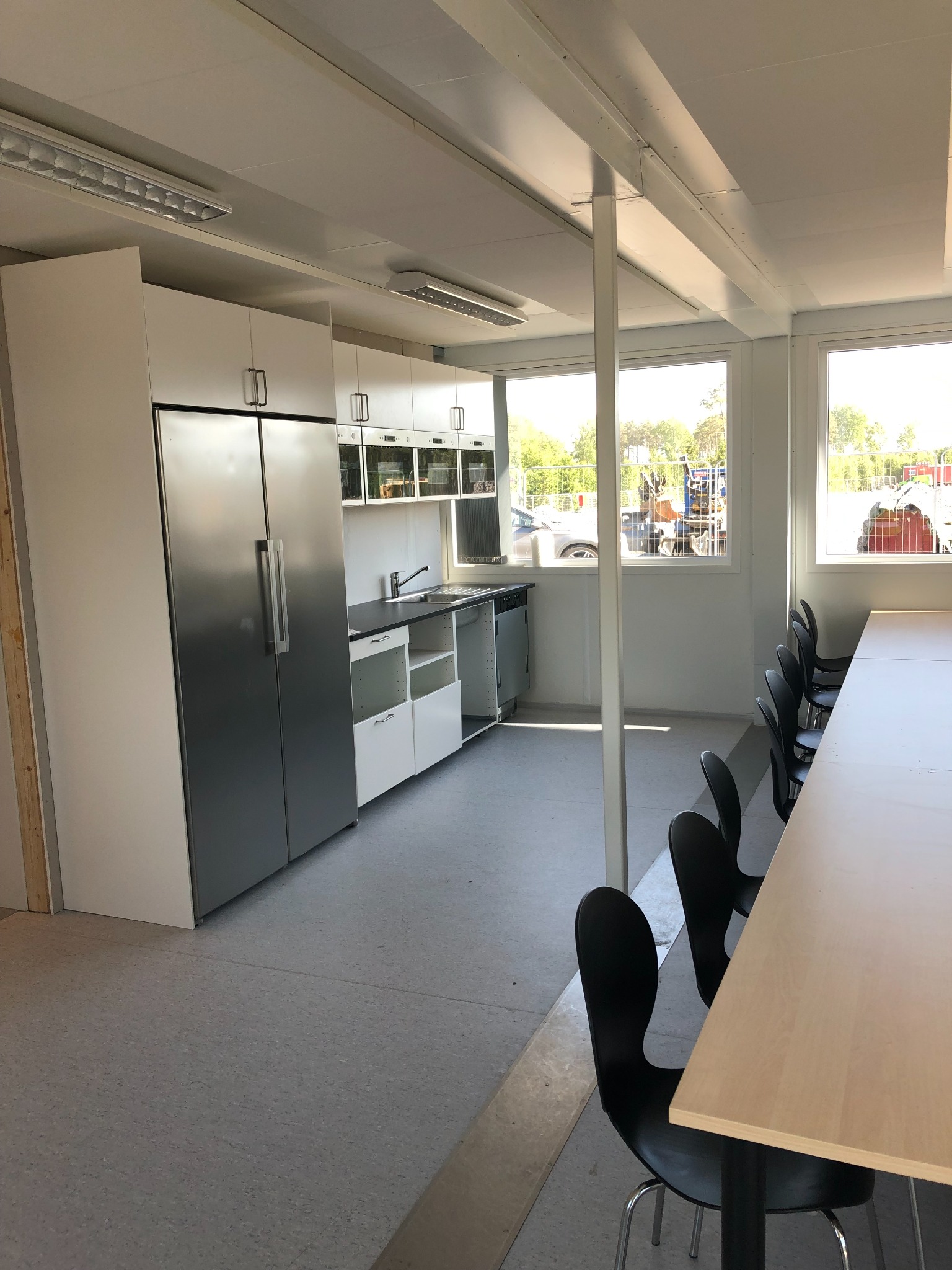 Wyposażona kuchnia biurowa z dużym, dwudrzwiowym lodówko-zamrażarką, białymi szafkami, czarnym blatem i rzędem krzeseł przy jasnym stole, widoczna przez okno budowa.