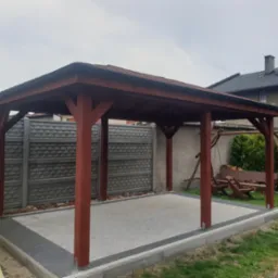 Drewniana altana ogrodowa z brązowymi słupami i dachem, wyłożona kostką brukową, w tle kościół i dom.