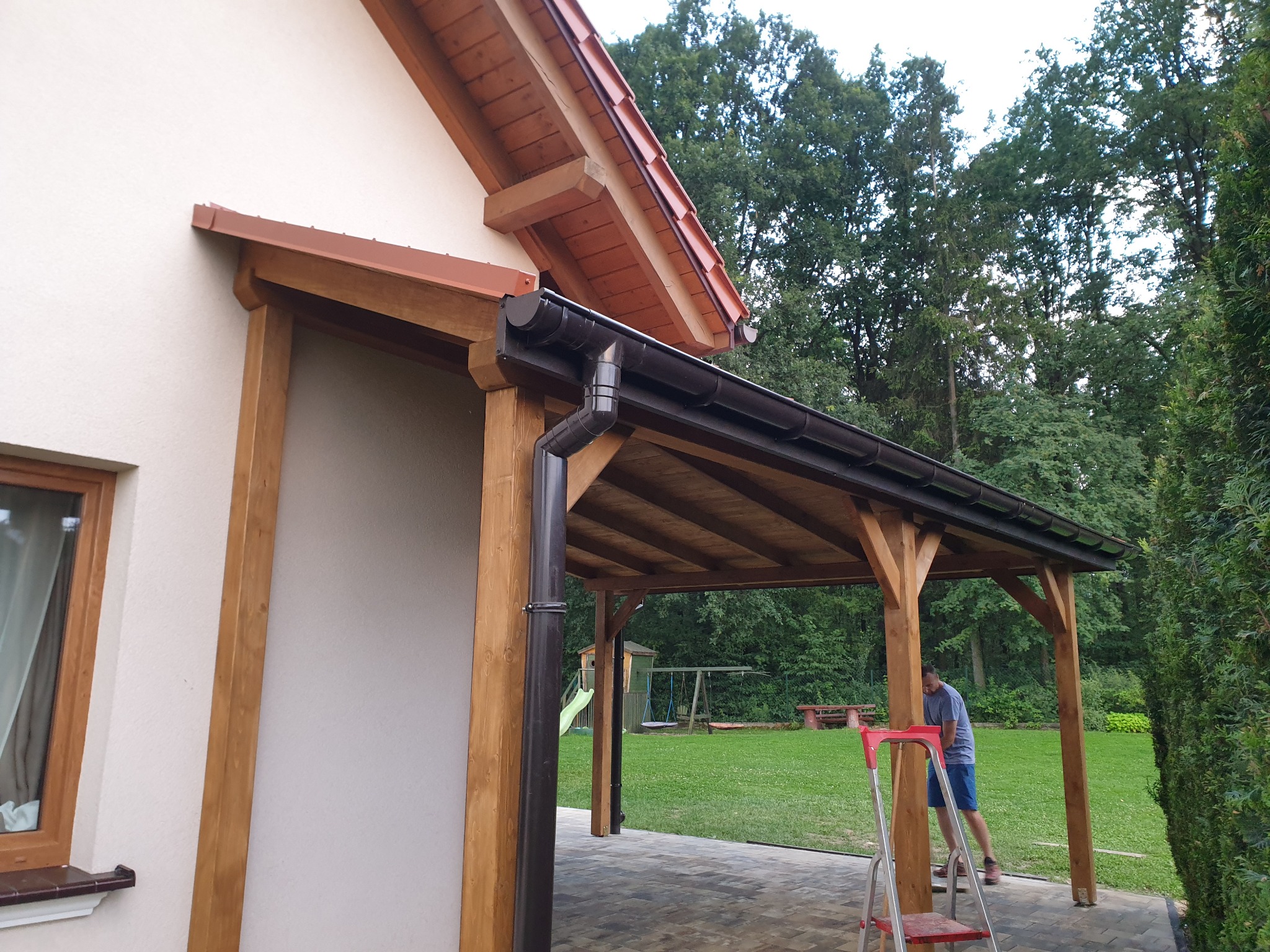 Drewniana konstrukcja zadaszenia tarasu przylegająca do ściany domu, widoczne rynny i pionowa rura spustowa, w tle zielony trawnik i plac zabaw dla dzieci, mężczyzna na drabinie wykonuje prace...