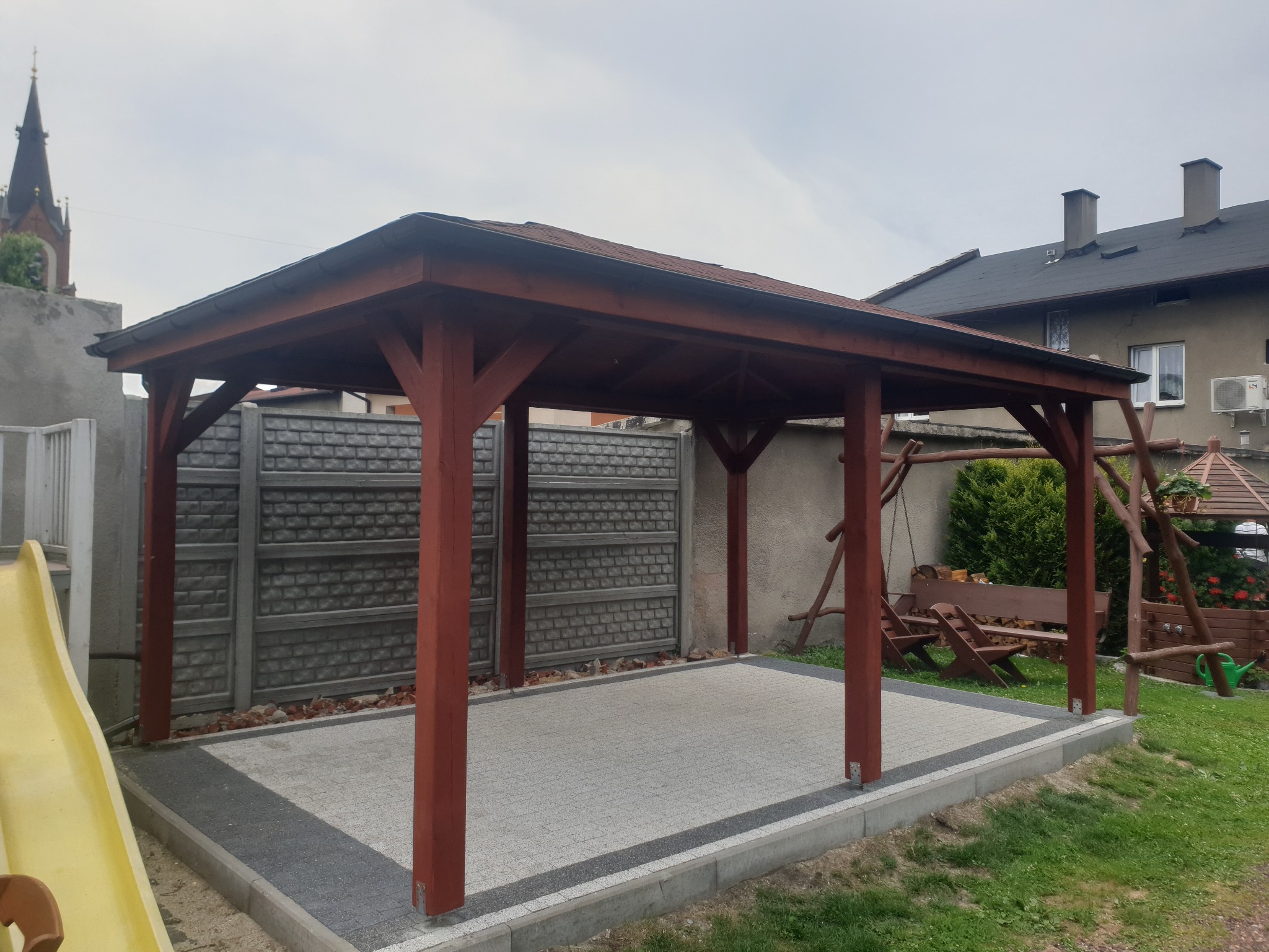 Drewniana altana ogrodowa z brązowymi słupami i dachem, wyłożona kostką brukową, w tle kościół i dom.